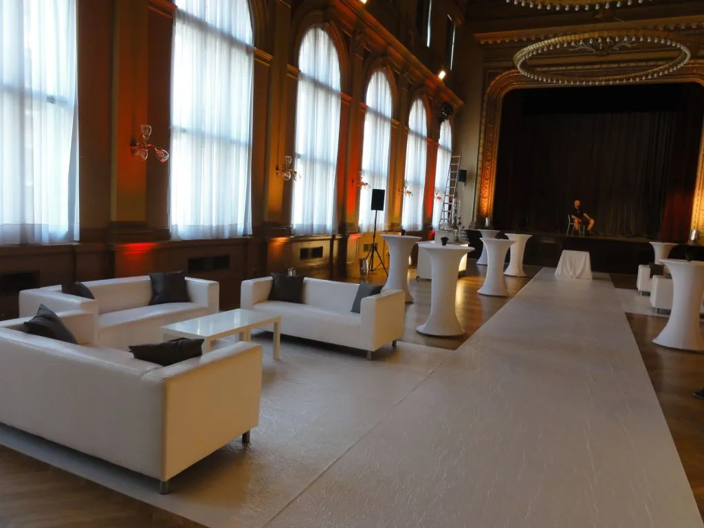 Interior of a grand hall with white sofas, high arched windows, and a long white carpet leading to a stage area