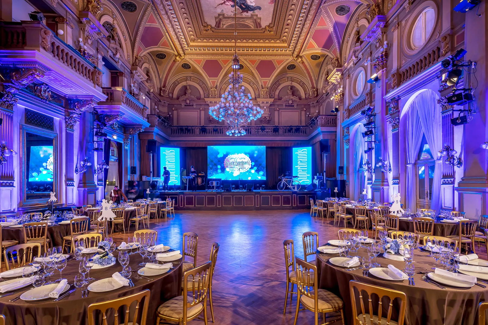 A grand hall with ornate ceilings, chandeliers, and round tables set for an event under purple lighting.