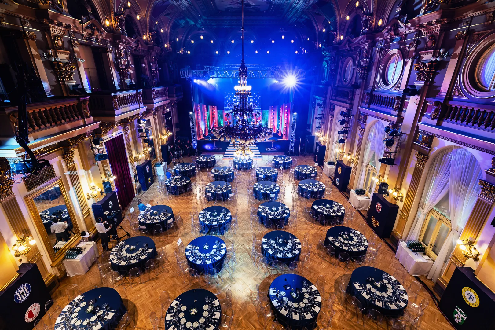 Ornate interior hall with high ceilings, chandeliers, and round tables arranged for an event under blue and purple lighting