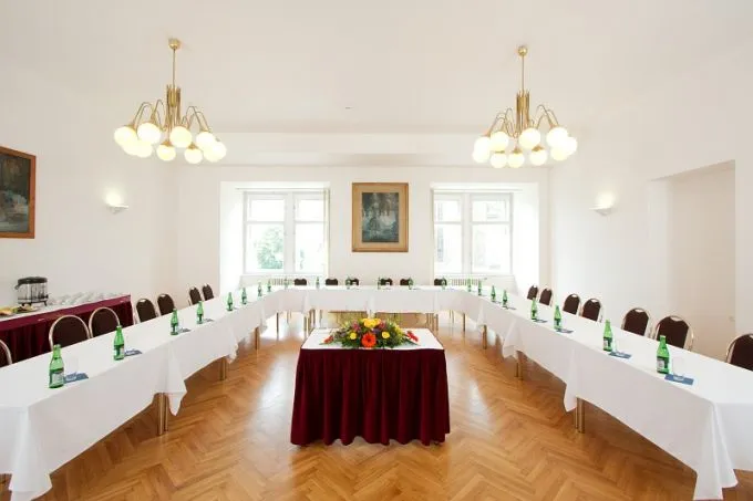 A spacious interior hall with wooden floors, white walls, and large windows, furnished with long tables covered in white cloths and a central table with a red cloth, under two chandeliers.