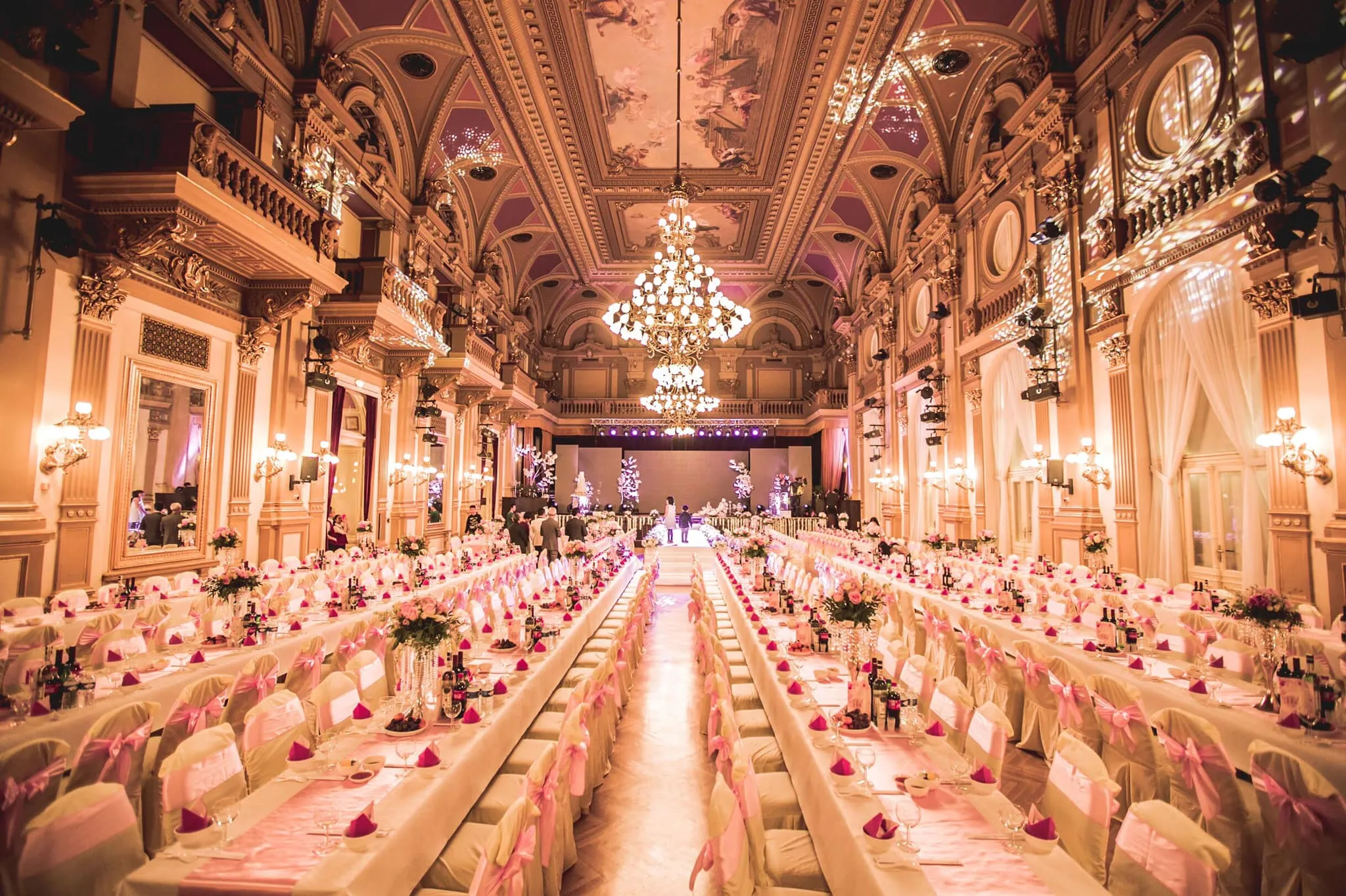 Ornate interior of a grand hall with long tables set for an event, chandeliers, and decorative ceilings