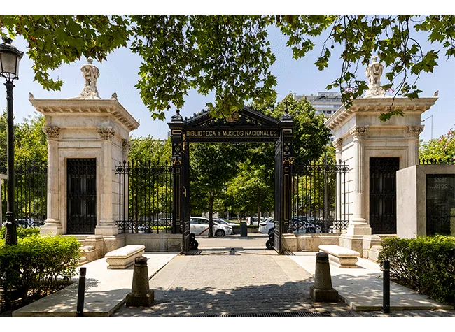 Kamienna brama z czarnymi żelaznymi wrotami i kamiennymi filarami przy wejściu do Museo de la Biblioteca Nacional, otoczona zielonymi drzewami i utwardzoną ścieżką.
