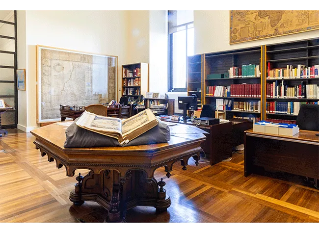 Wnętrze biblioteki z dużym drewnianym stołem, otwartą księgą, regałami i mapą na ścianie