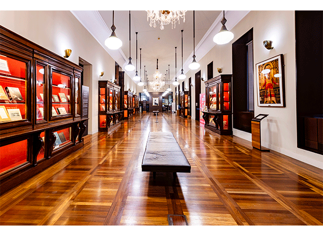 Długa galeria z drewnianymi podłogami, gablotami i żyrandolami w Museo de la Biblioteca Nacional