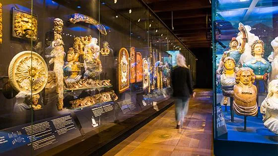 Interior gallery with glass display cases showing gilded maritime artifacts and ship figureheads, with a visitor walking through