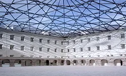 Courtyard with geometric glass and steel roof structure, white building walls with arched ground-floor openings and multiple windows
