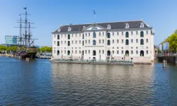 White historic building with dark roof along a canal, featuring a tall-masted ship docked nearby under a clear blue sky