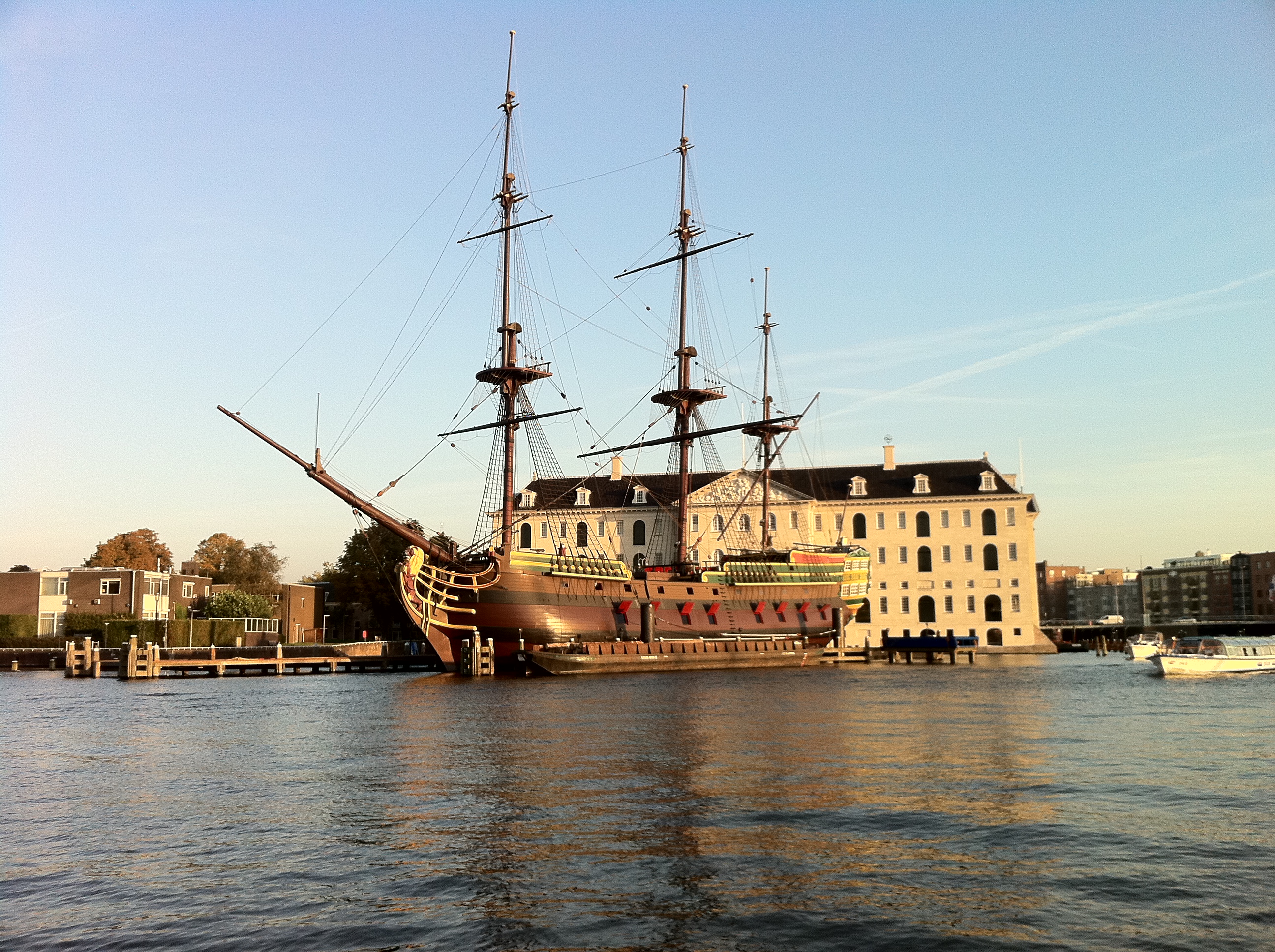 A large historic ship docked in front of a white building with a dark roof, situated on a waterway under a clear sky