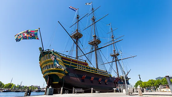 Historic three-masted ship with colorful flags docked at the National Maritime Museum, featuring ornate bow and Dutch flags under a clear blue sky