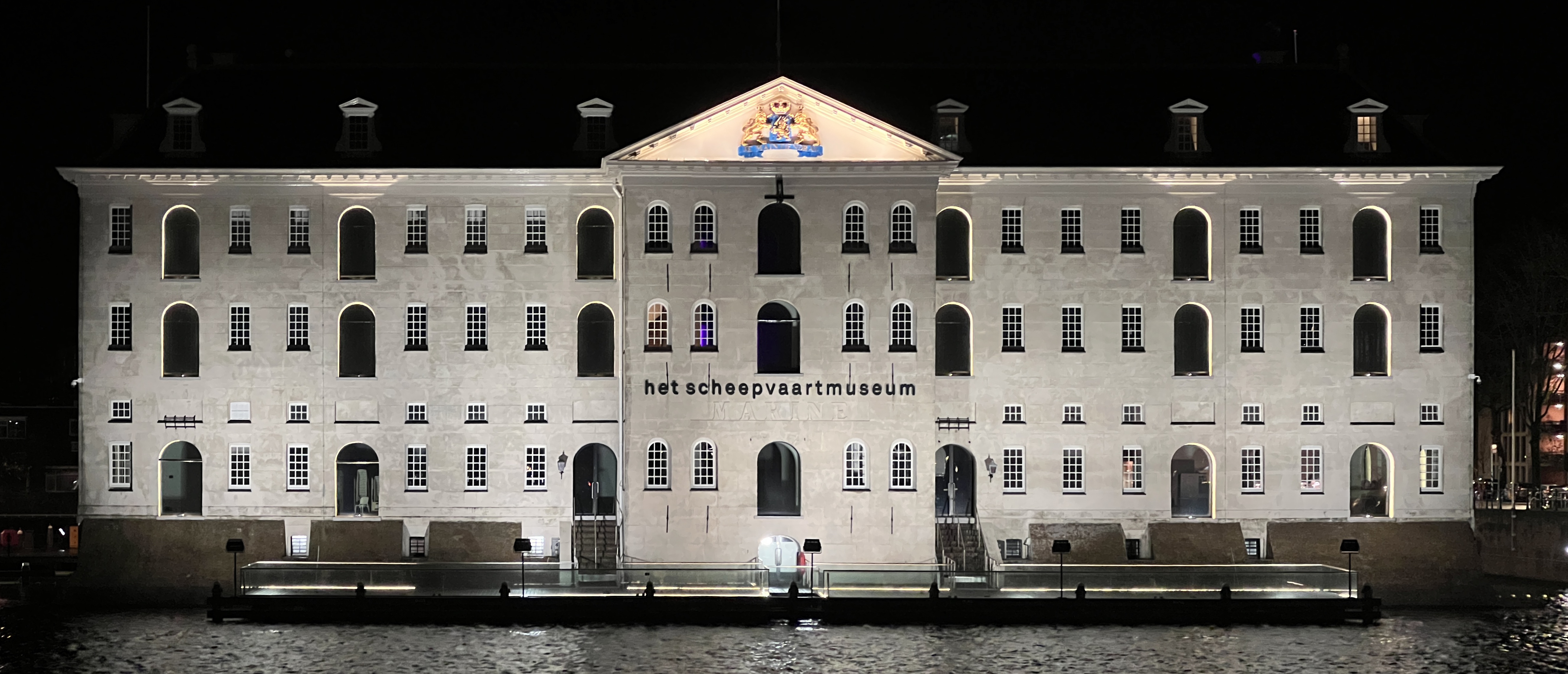 Nighttime view of the illuminated National Maritime Museum facade with 'Het Scheepvaartmuseum' text visible on the building and water in the foreground.