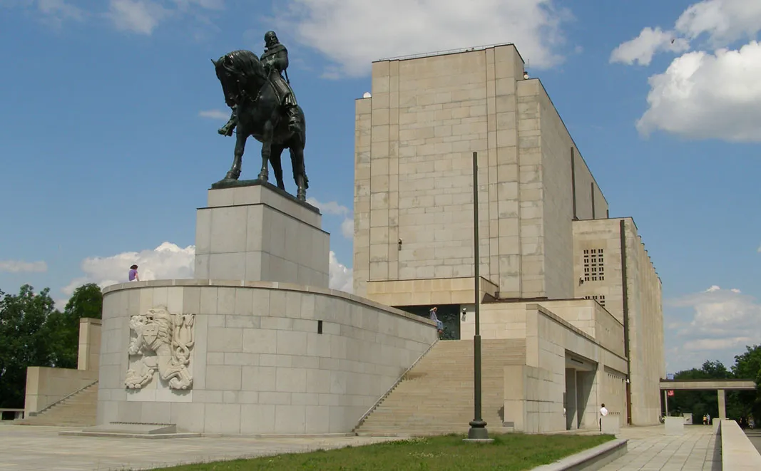 Una gran estatua ecuestre sobre un pedestal de piedra junto a un edificio modernista de hormigón bajo un cielo azul con nubes