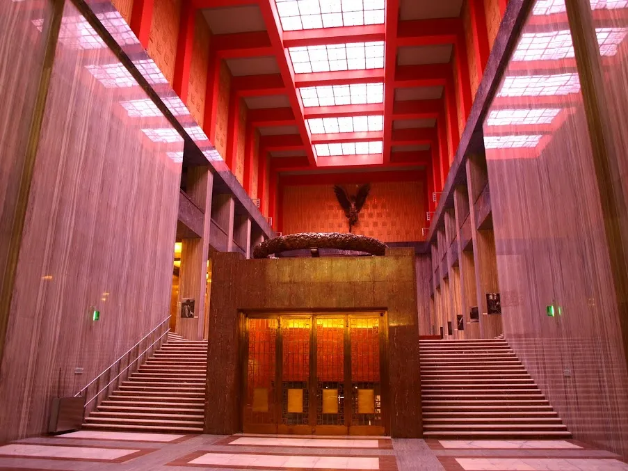 Interior de un gran salón con vigas rojas, una entrada central dorada y una gran estatua ecuestre a lo lejos.