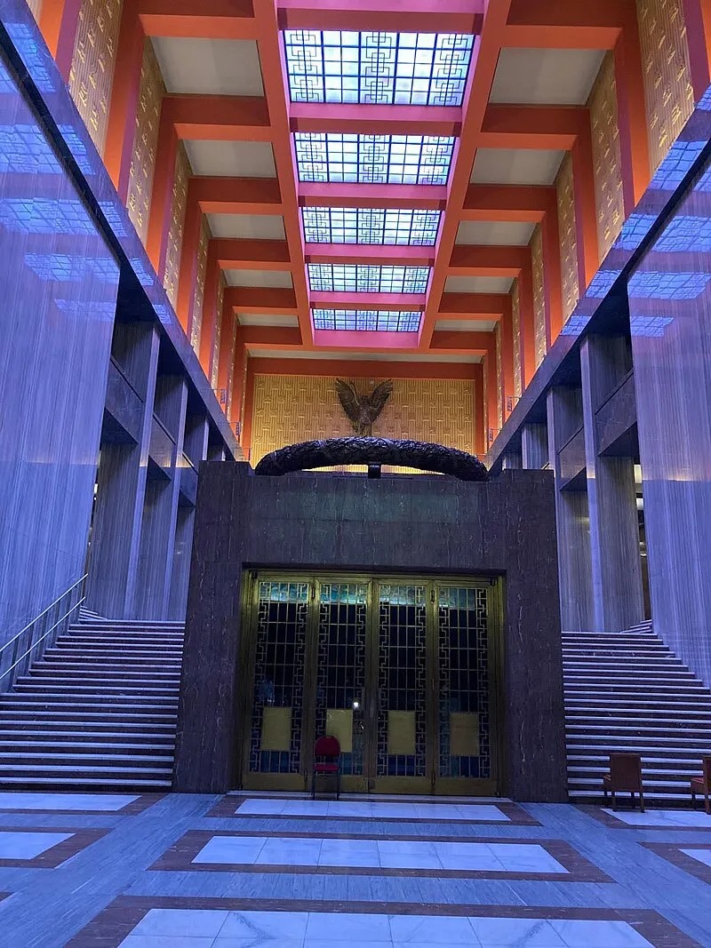 Sala interior con vigas rojas, tragaluces y una entrada central con una escultura de bronce sobre las puertas