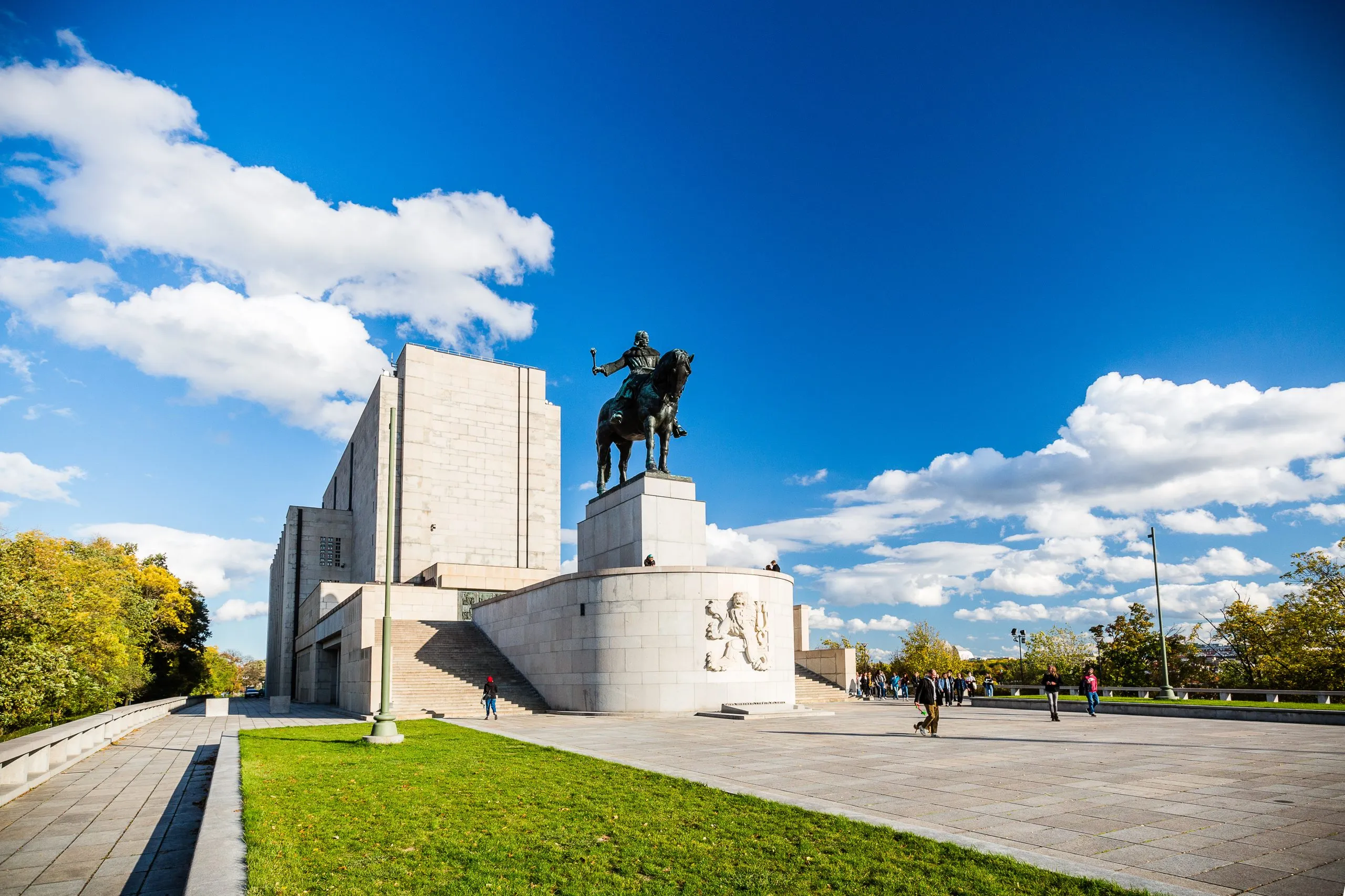 Una gran estatua ecuestre sobre un pedestal frente a un edificio modernista en la colina Vítkov, con hierba verde y un cielo azul.