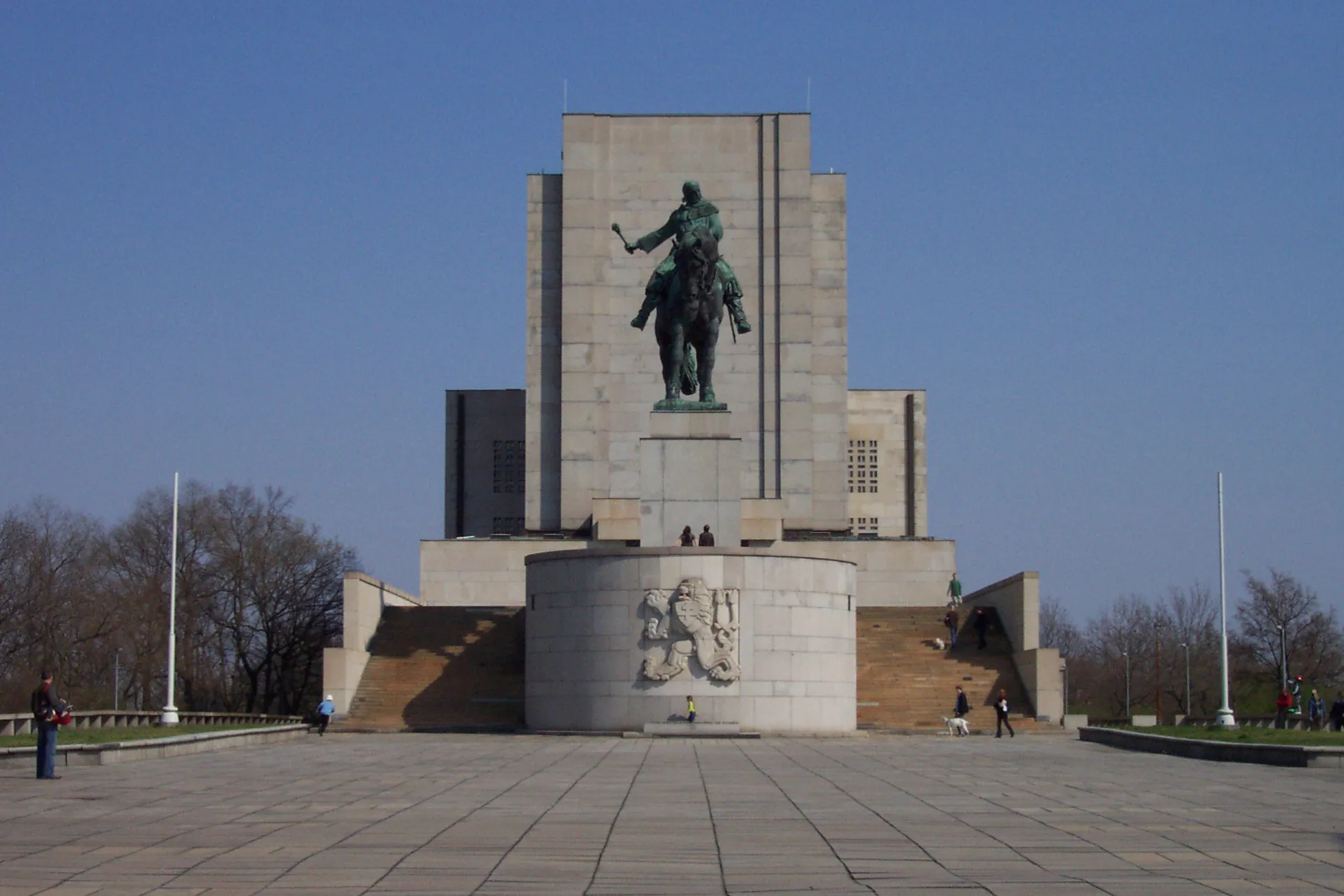 Un gran monumento de hormigón con una estatua ecuestre verde en la parte superior, contra un cielo azul claro, con una plaza pavimentada en primer plano.