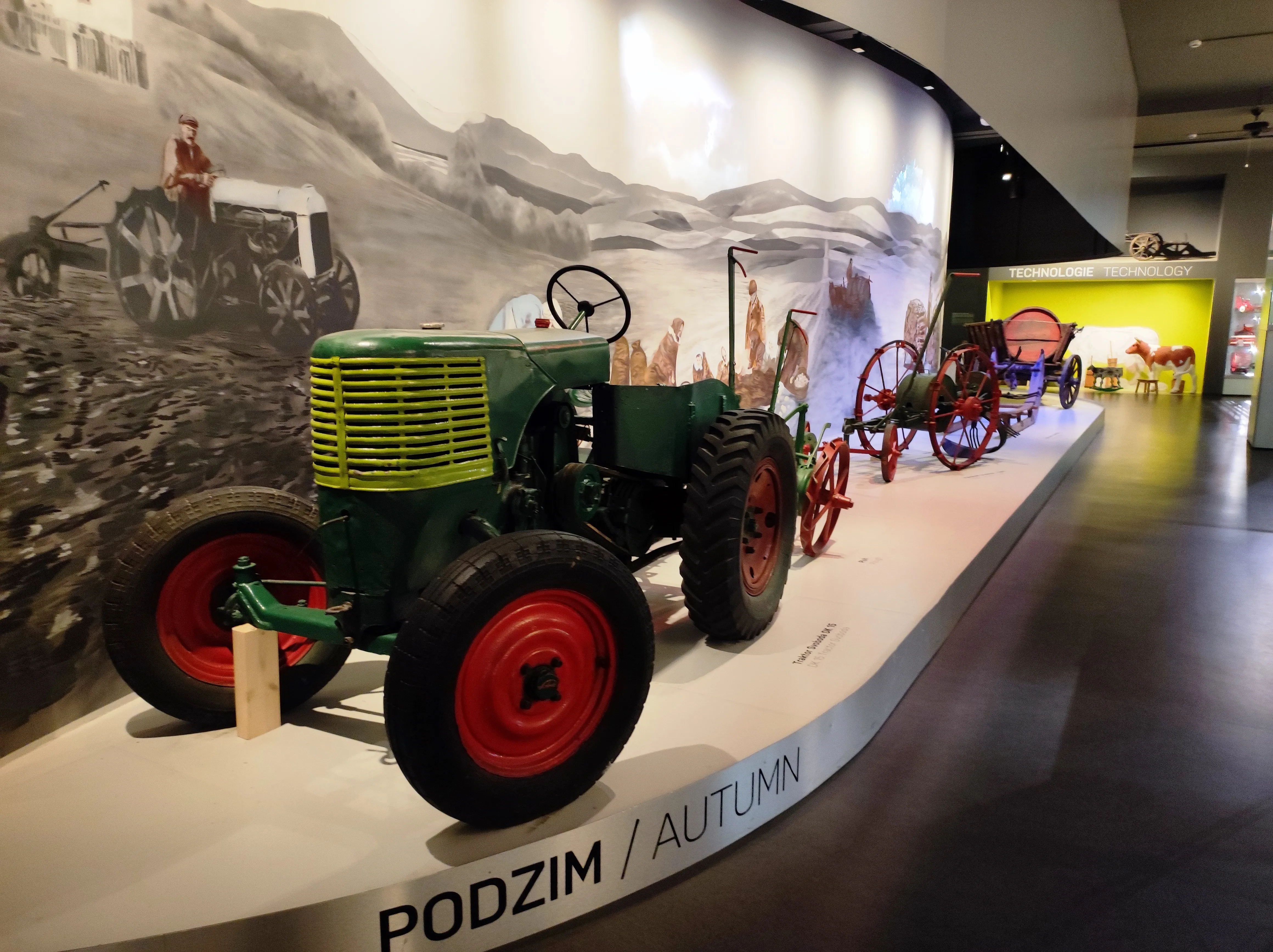 A green tractor with red wheels on display in a museum exhibit with a mural of rural life in the background