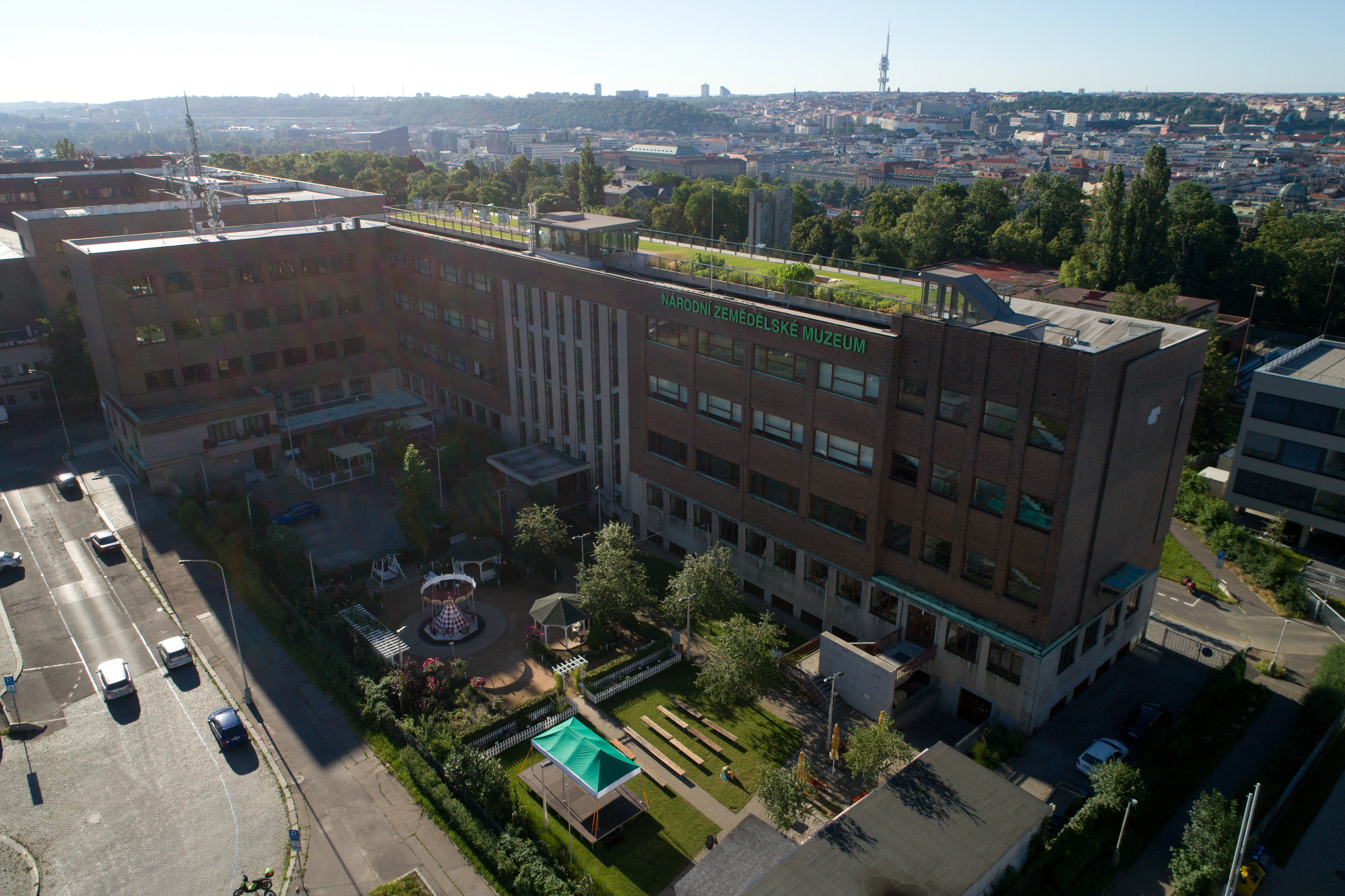 Aerial view of a large brick building with a green roof, surrounded by trees and a paved area with cars, with a cityscape in the background.