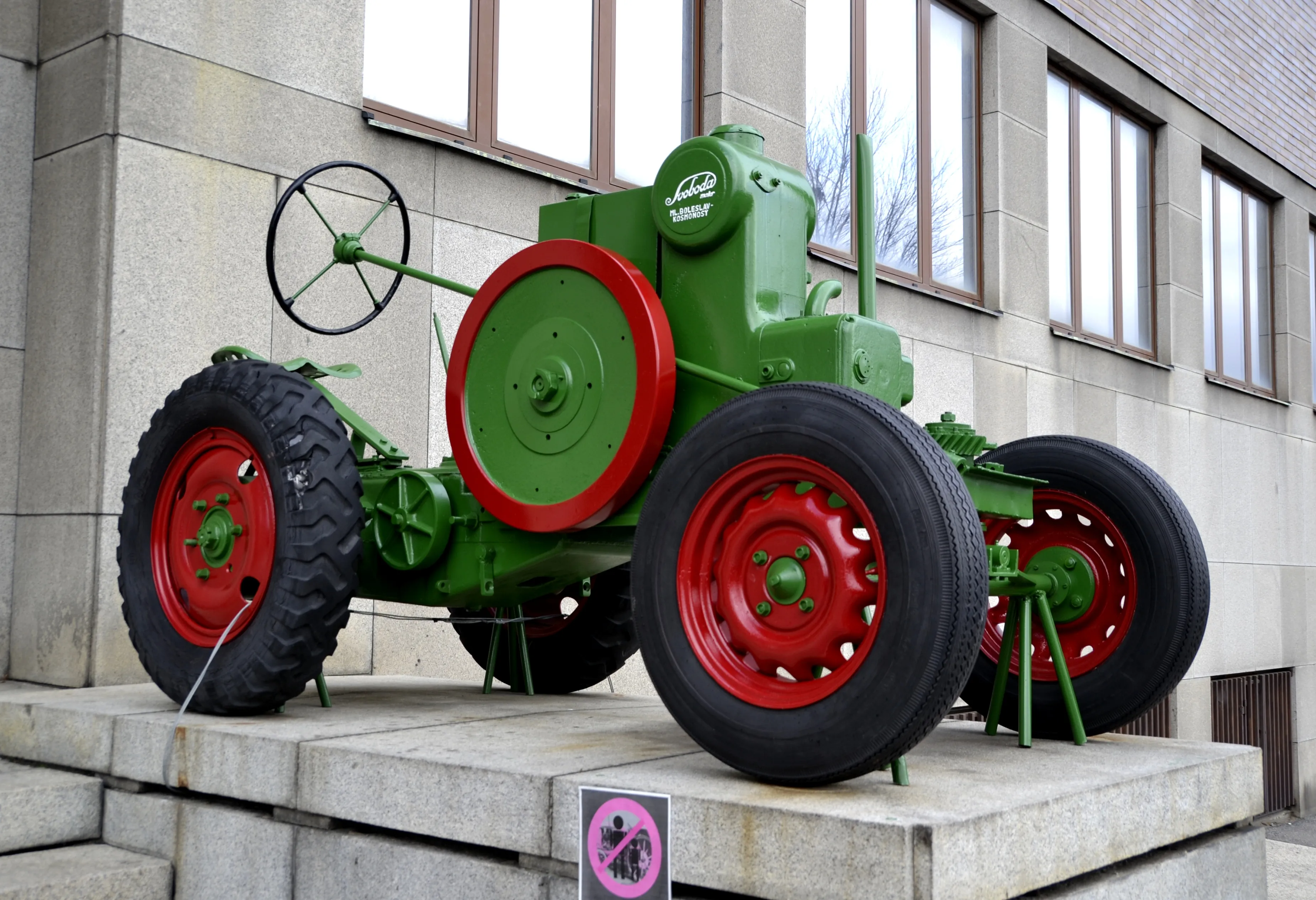 A green vintage tractor with red wheels on a stone pedestal in front of a building