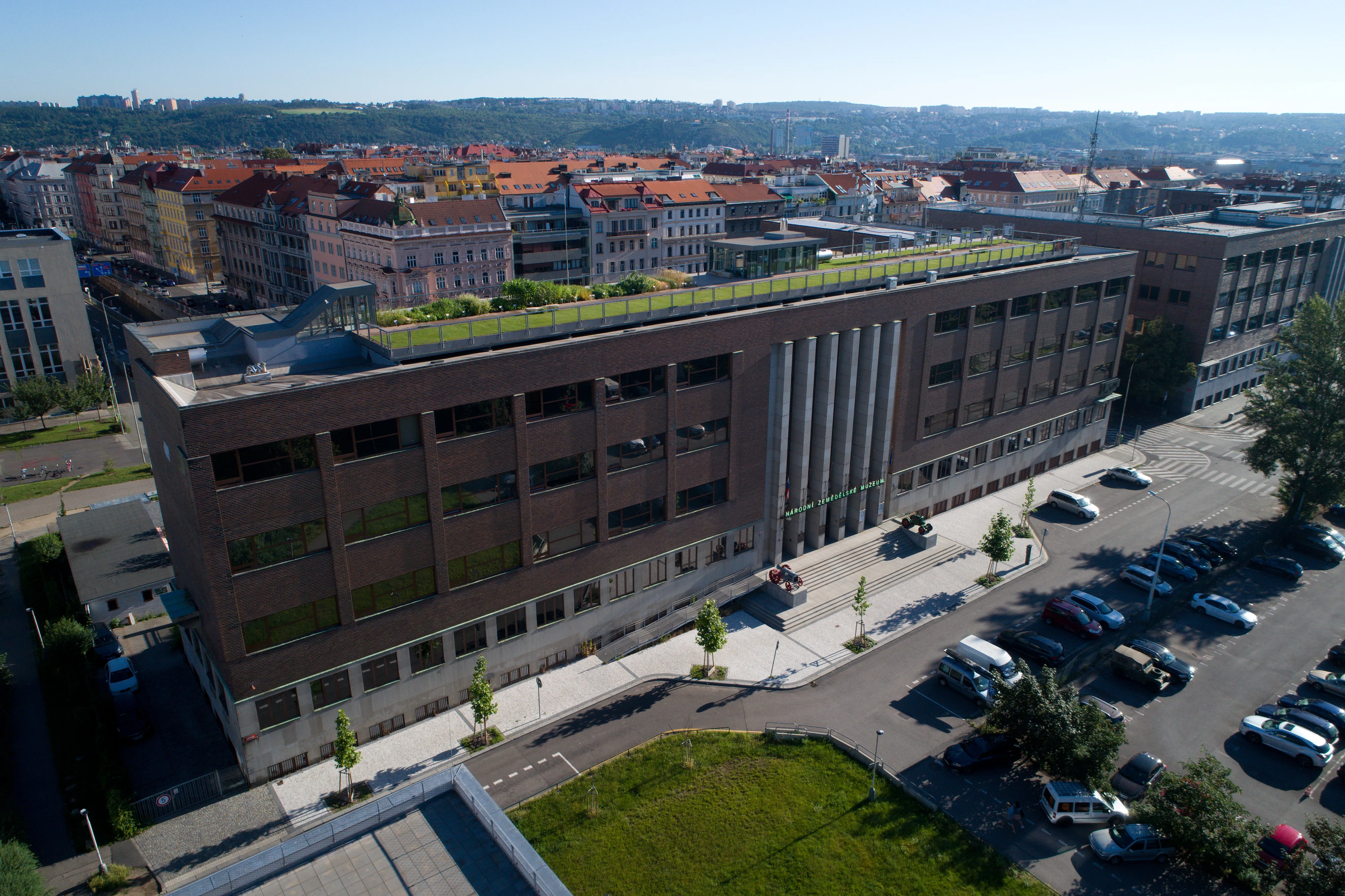 Aerial view of a modern building with a green roof, surrounded by parked cars and city buildings.