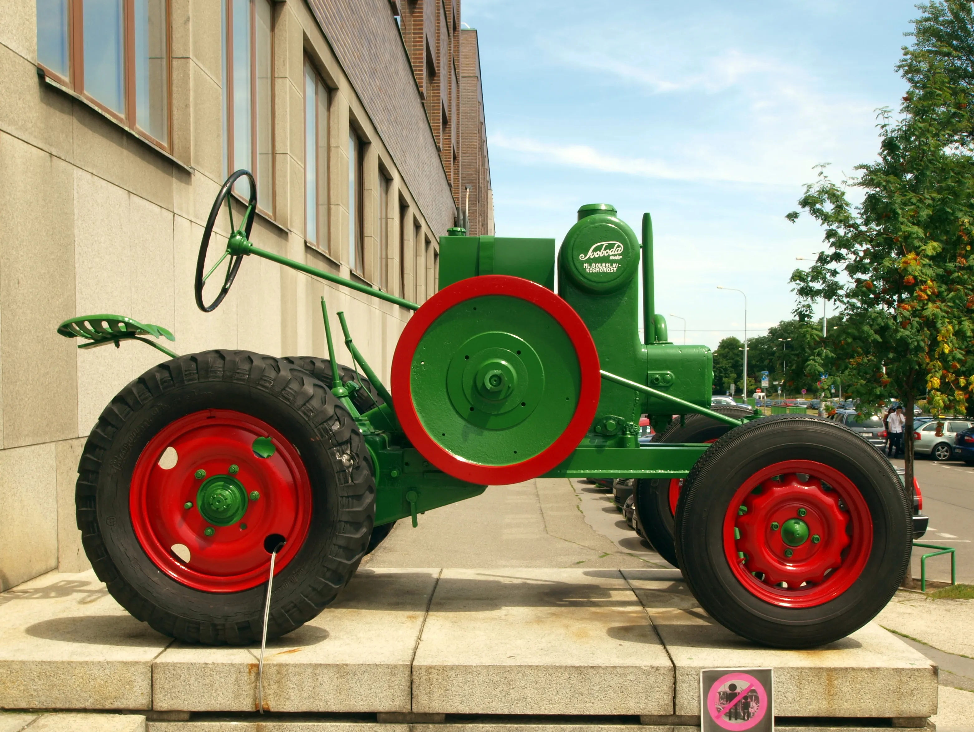 A green vintage tractor with red wheels and a large red wheel cover displayed on a stone pedestal outside a building.