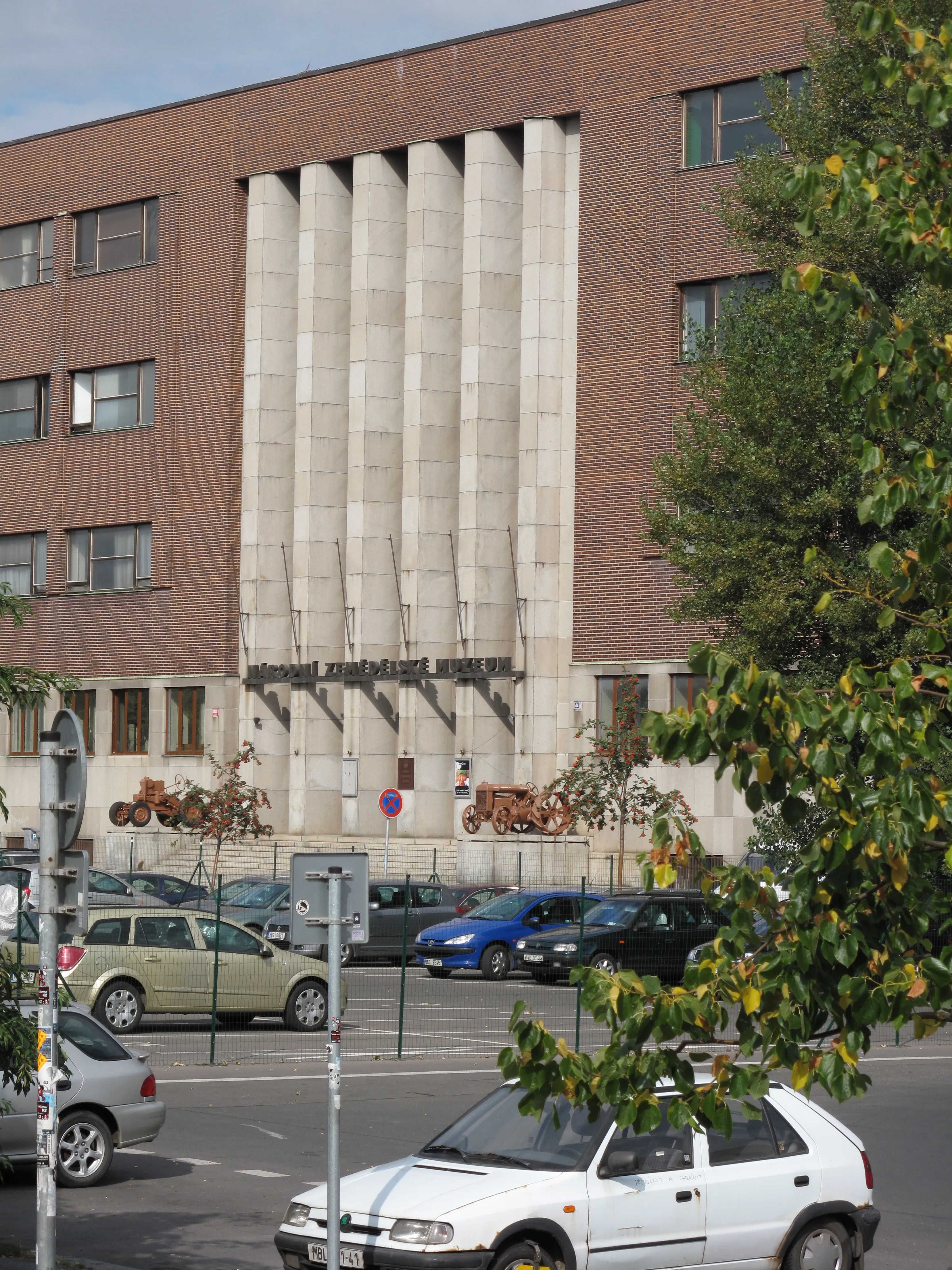 Bakstenen en betonnen gebouw met verticale kolommen en een bord met de tekst 'National Museum of Agriculture', omgeven door bomen en geparkeerde auto's