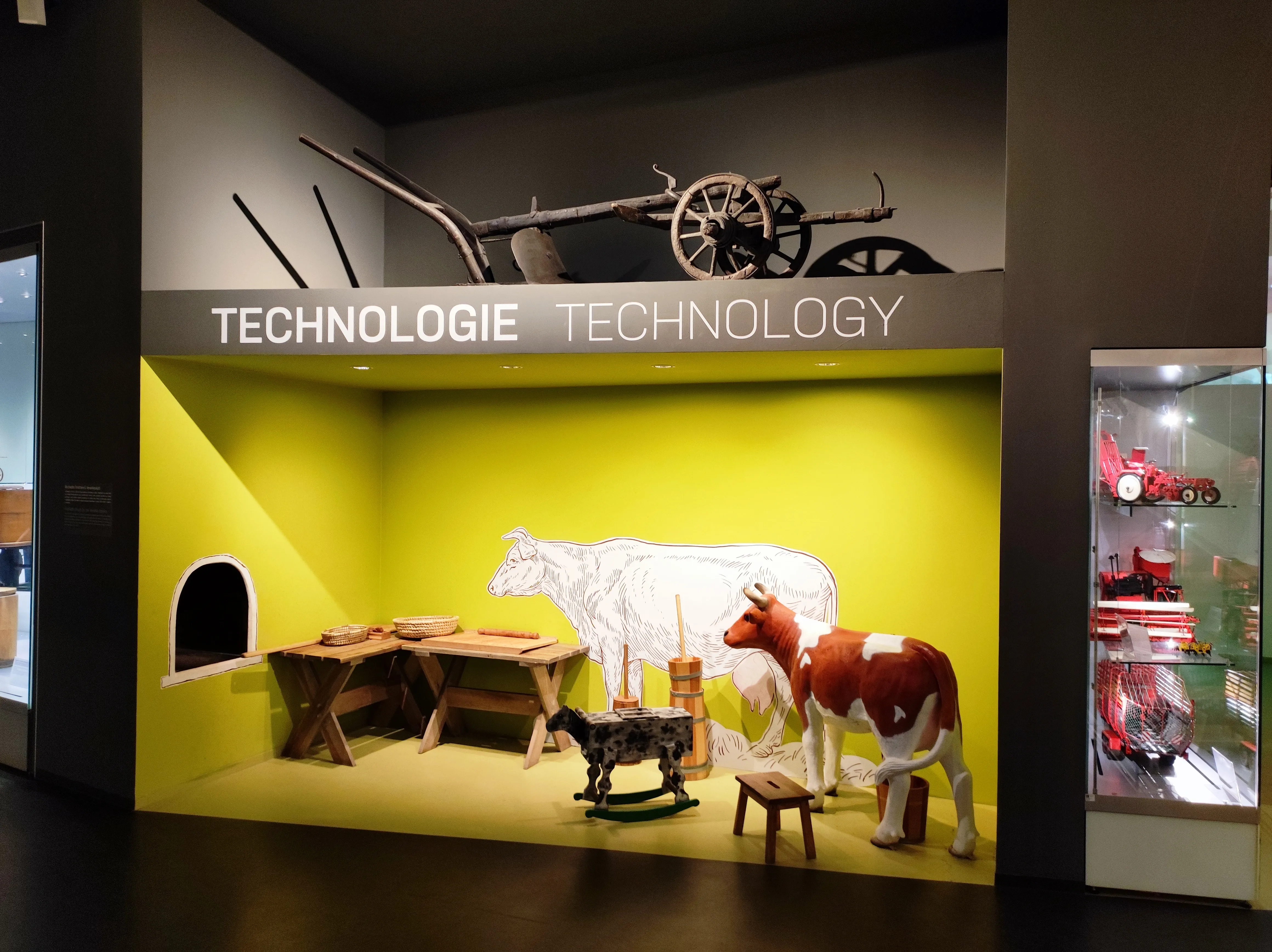 Exhibition display with a yellow wall, a wooden cart, a cow model, and a sign reading TECHNOLOGIE TECHNOLOGY