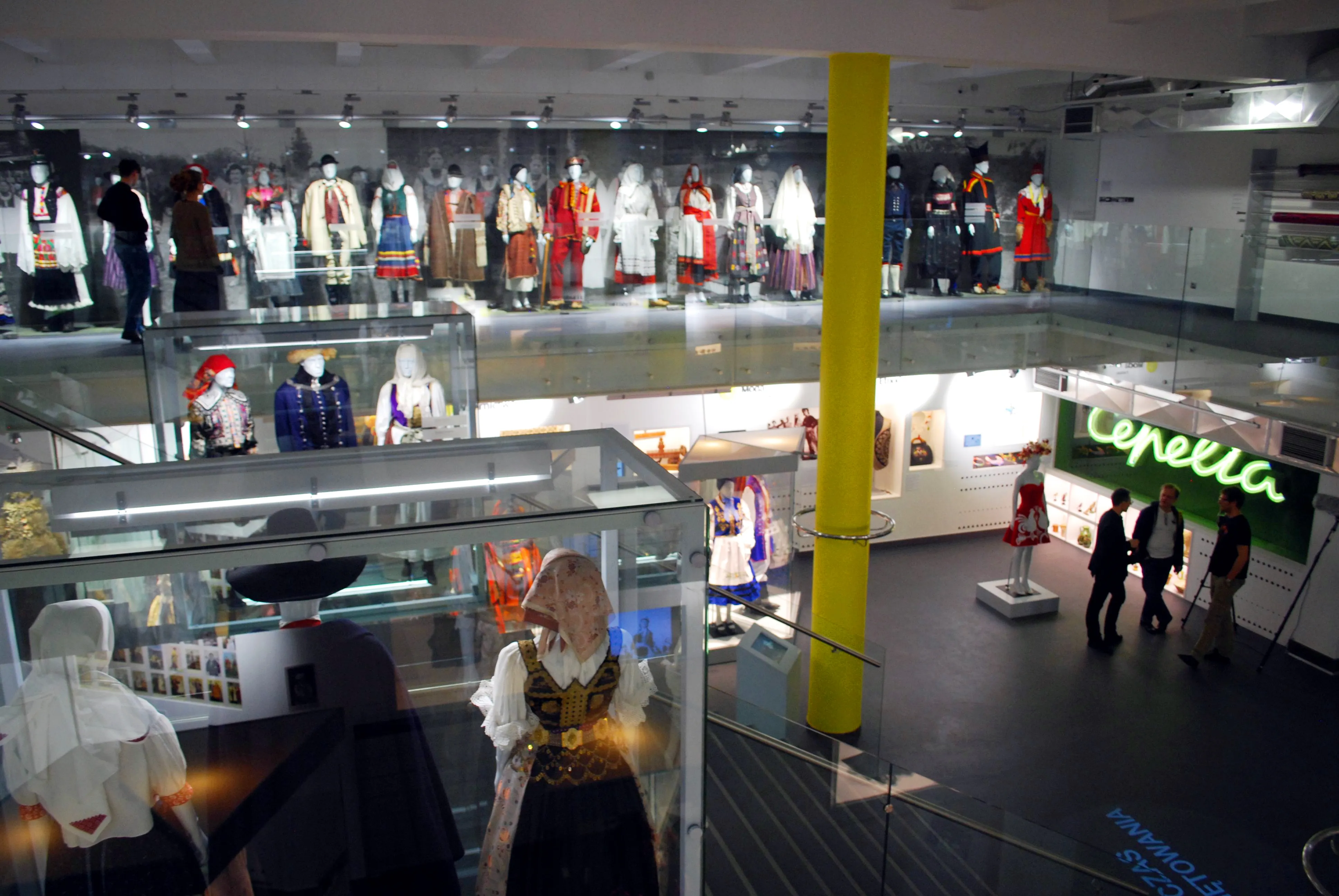 Espacio de exposición con trajes tradicionales en maniquíes, un pilar amarillo y un letrero de neón