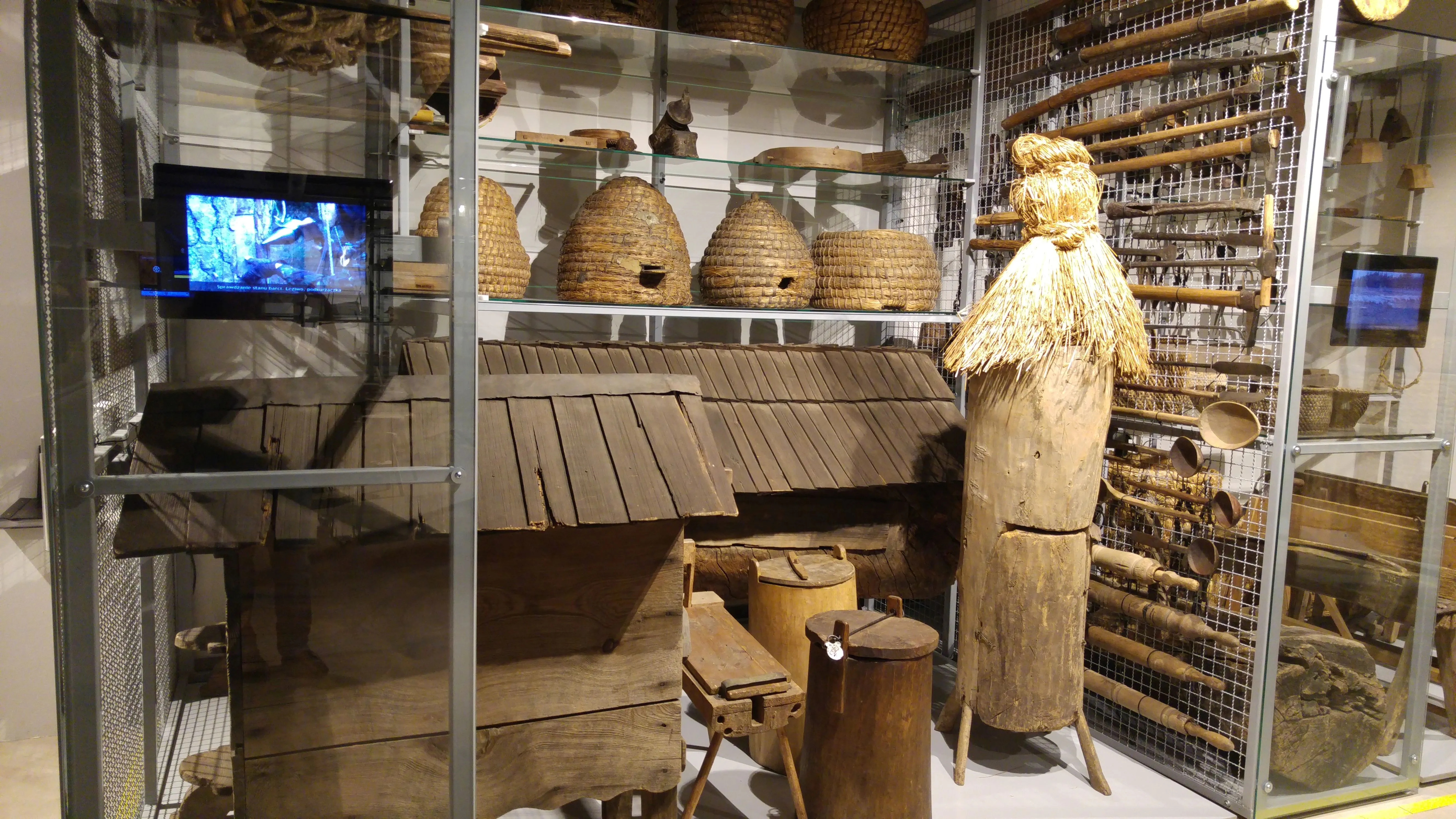 Exposición de estructuras tradicionales de madera, colmenas y una figura de madera tallada en una vitrina