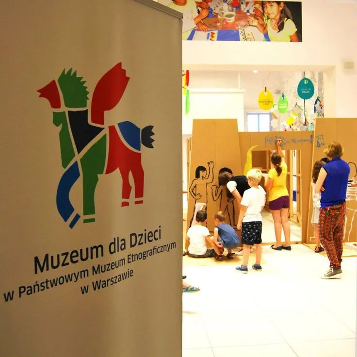 Un cartel para el Museo de Niños en el Museo Nacional de Etnografía de Varsovia, con un colorido logo de caballo y texto, con niños y adultos en un área de exposición detrás.
