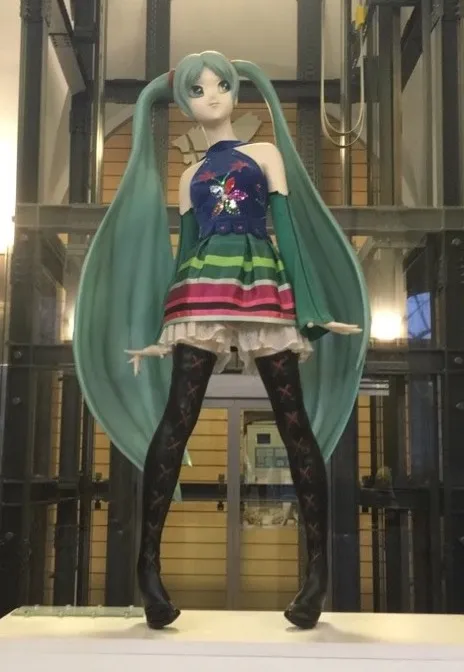 Una figura grande de un personaje con cabello color turquesa y un vestido colorido de pie en una vitrina