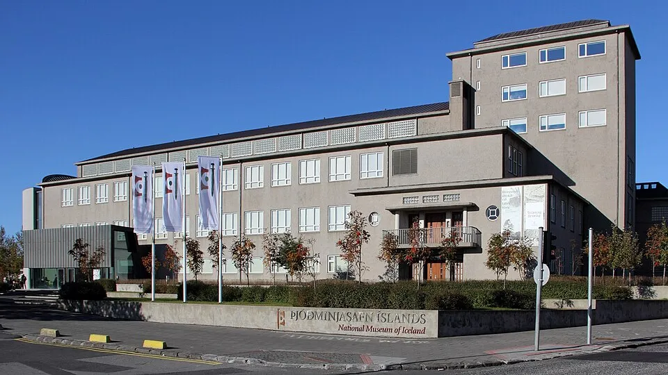 Szeroki widok z zewnątrz na budynek Muzeum Narodowego Islandii, wielopiętrową betonową konstrukcję z pionowymi banerami i niskim szyldem pod czystym błękitnym niebem.