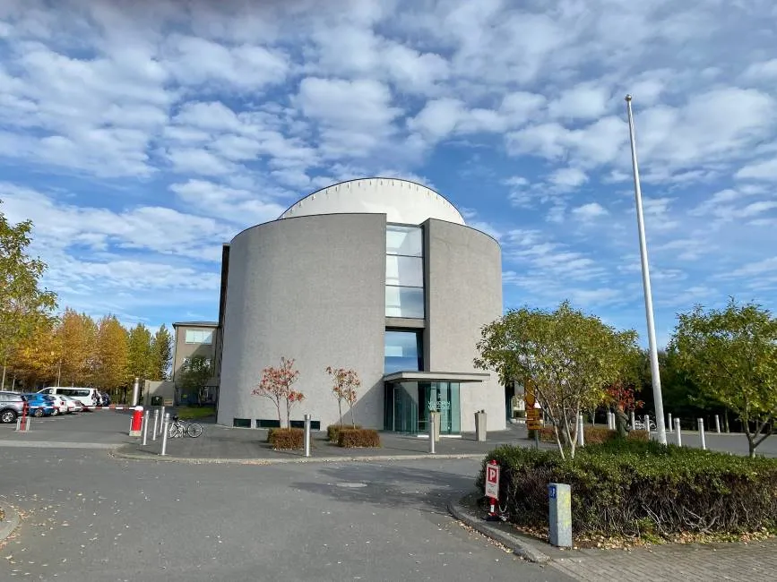 Widok z zewnątrz na Muzeum Narodowe Islandii, ukazujący charakterystyczny cylindryczny budynek z białą kopułą, szklaną fasadą wejściową, otoczony jesiennymi drzewami i parkingiem pod częściowo zachmurzonym niebieskim niebem