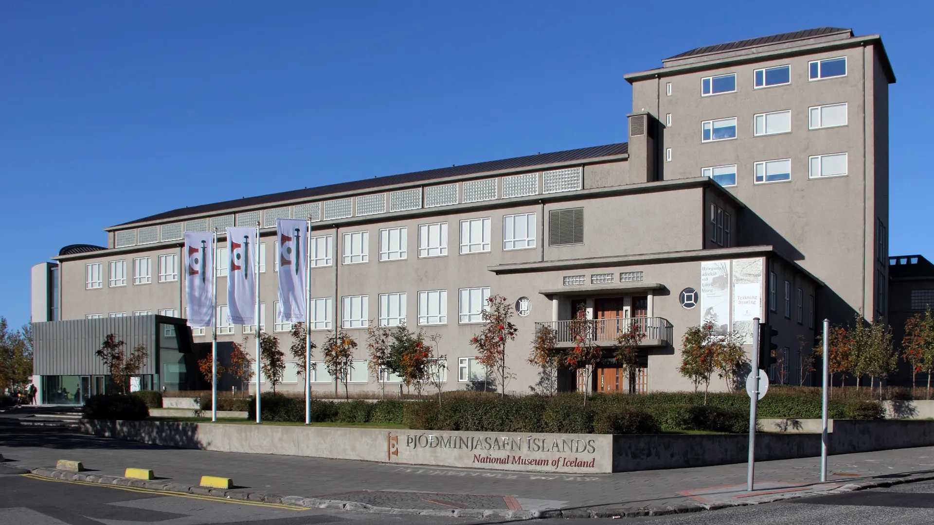 Widok z zewnątrz na budynek National Museum of Iceland ze znakiem przed nim i flagami zawieszonymi na fasadzie pod bezchmurnym, niebieskim niebem.