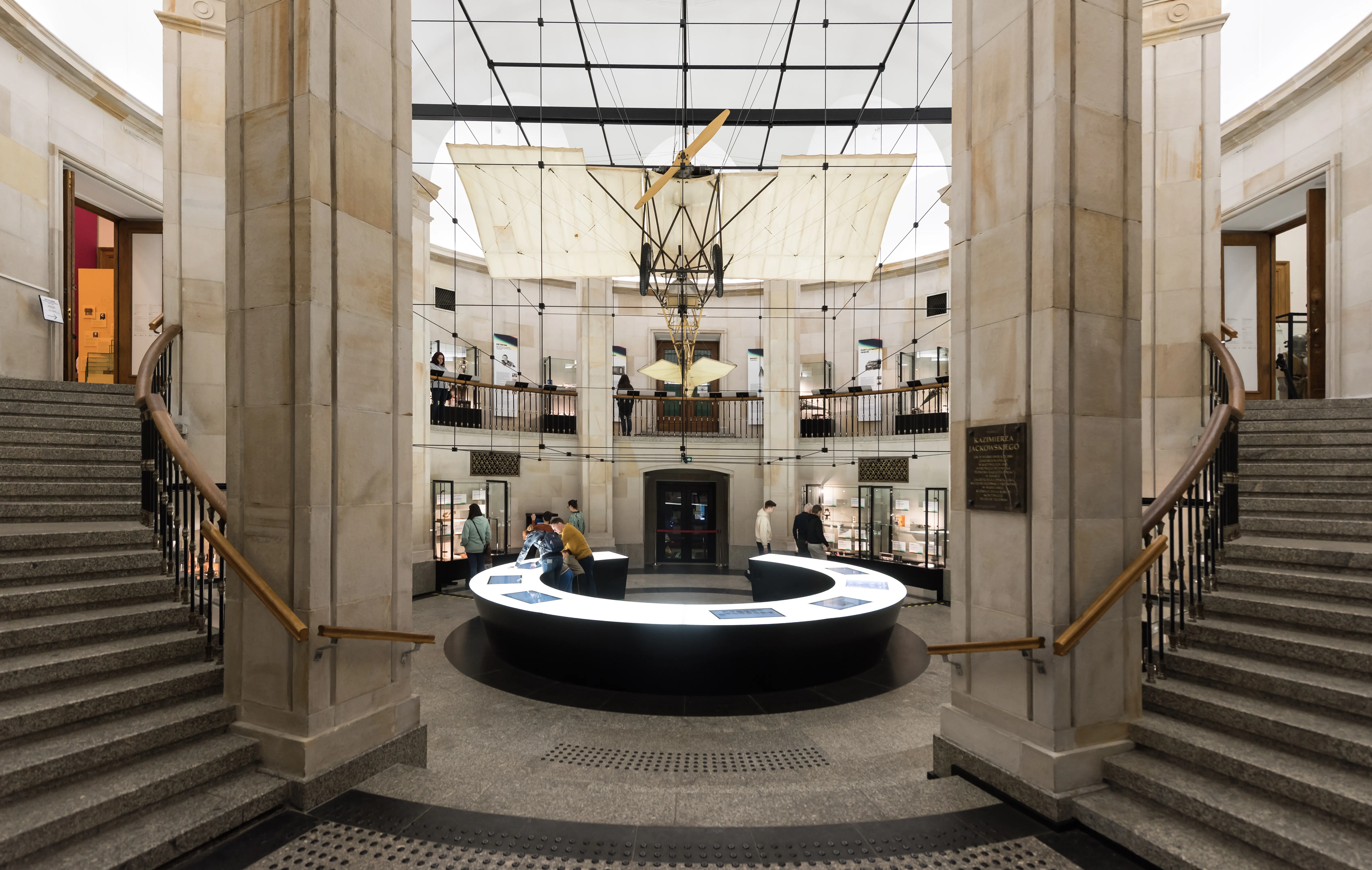 Gran vestíbulo interior con columnas de piedra, dos escaleras y una exposición circular central bajo un techo de cristal