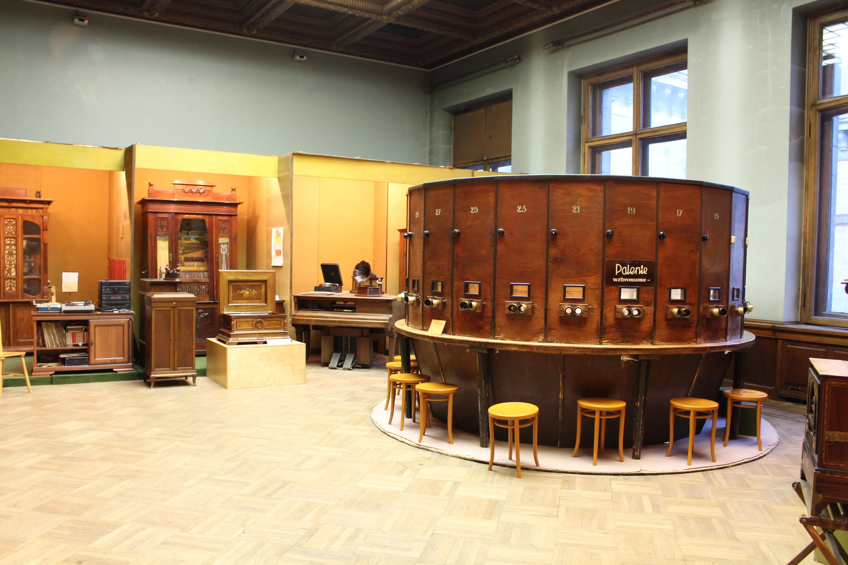 Interior de un museo con una gran exposición circular de madera que contiene cabinas telefónicas antiguas, rodeada de taburetes de madera, con otros artefactos históricos y vitrinas al fondo.