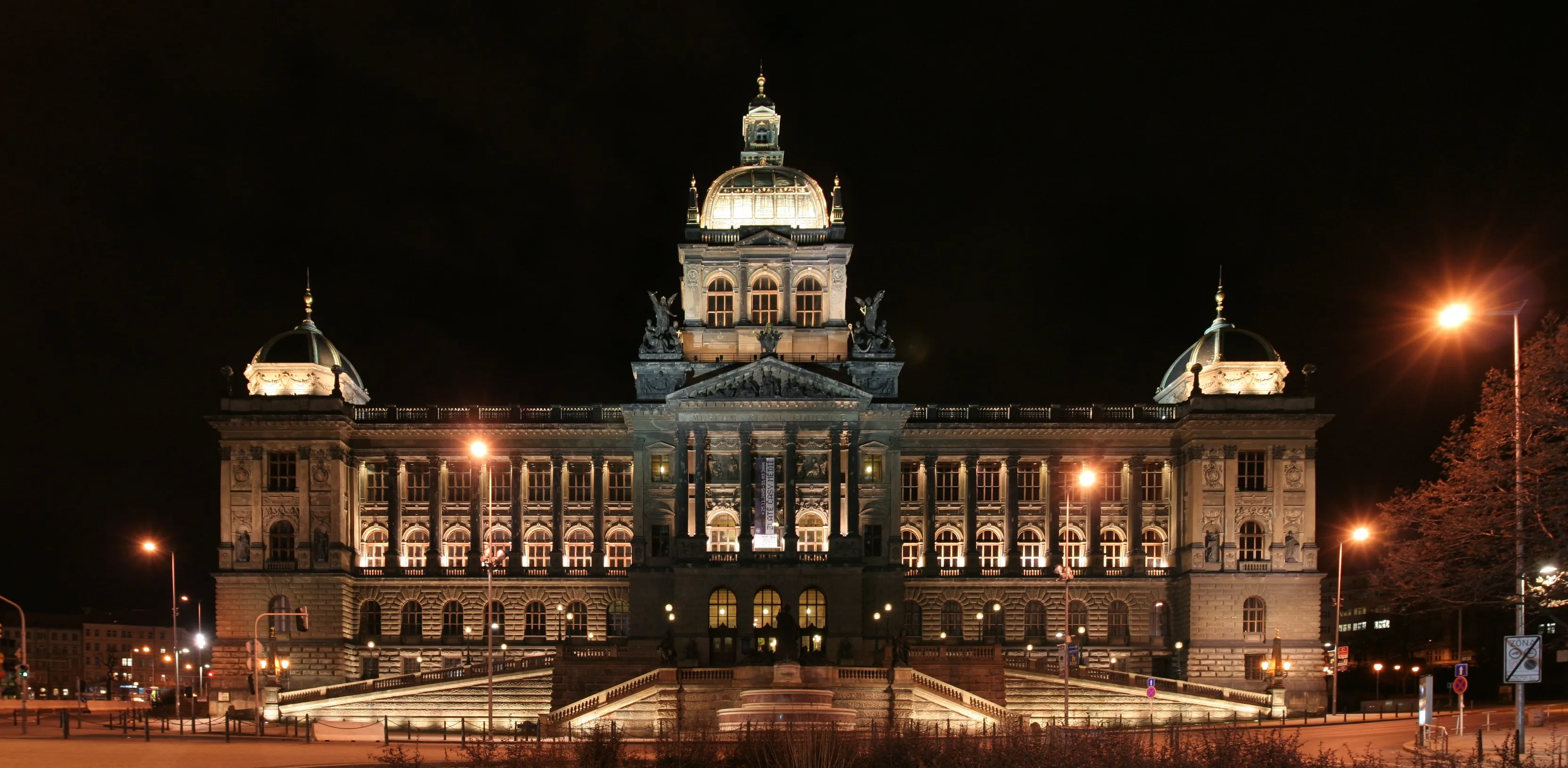 Neoclassicistisch gebouw met een centrale koepel en twee zijkoepels, 's nachts verlicht, met straatverlichting en bomen op de voorgrond.