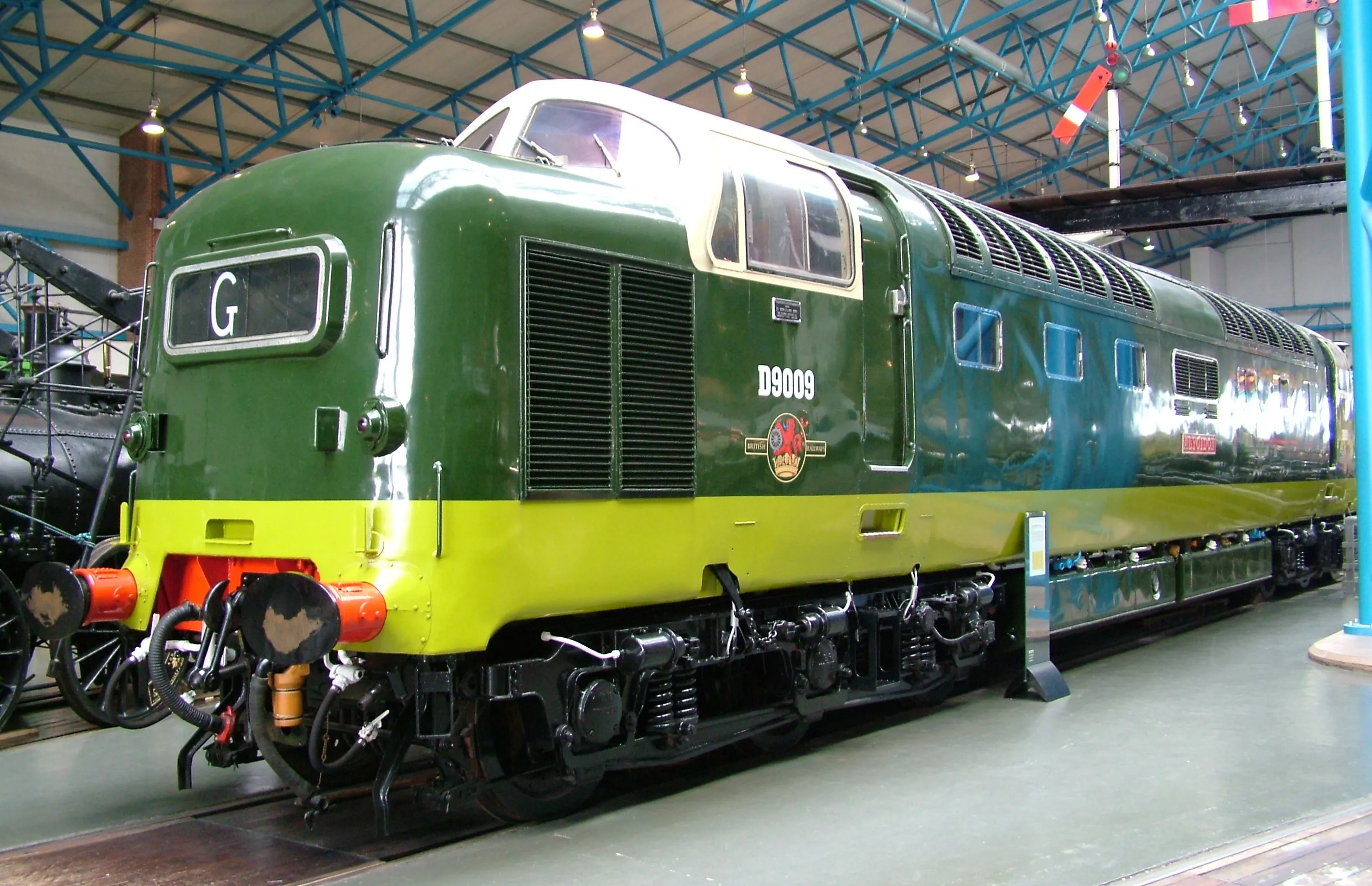Zielono-żółta lokomotywa spalinowa Deltic D9009 Alycidon wystawiona w hali muzealnej z niebieskimi stalowymi dźwigarami dachowymi i oświetleniem górnym.