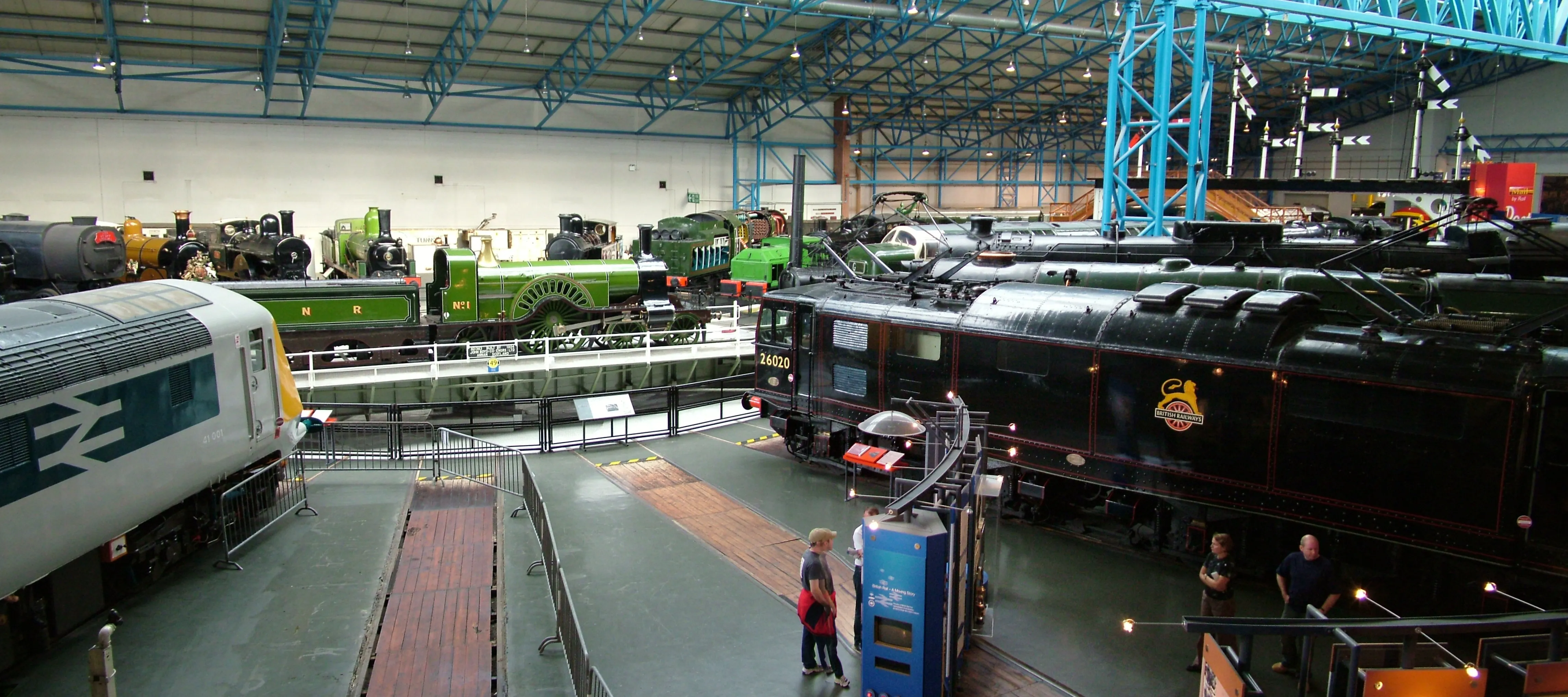 Szeroki widok na wnętrze Great Hall w National Railway Museum z wieloma zabytkowymi lokomotywami, w tym dużą czarną lokomotywą parową i pociągiem British Rail, oraz zwiedzającymi przechadzającymi się po parterze pod niebieską stalową konstrukcją dachu.