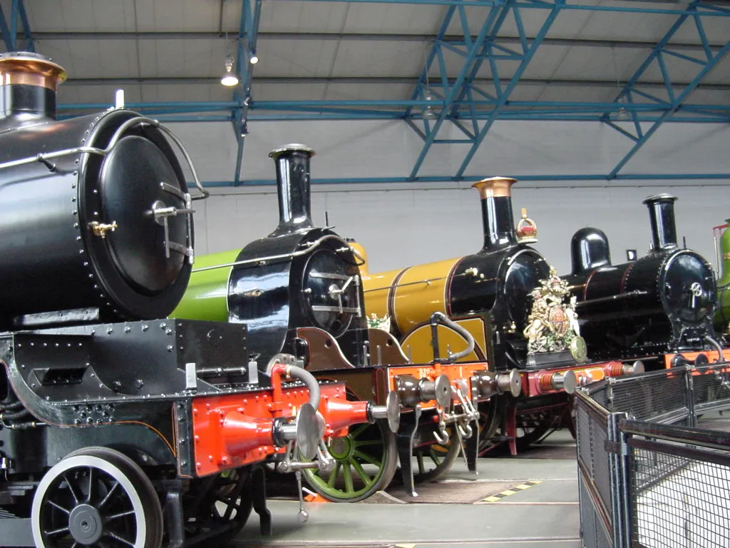 Szereg zabytkowych lokomotyw parowych w czarnym, zielonym i żółtym malowaniu, prezentowanych wewnątrz wielkiej hali wystawowej National Railway Museum ze stalowym sufitem kratownicowym.