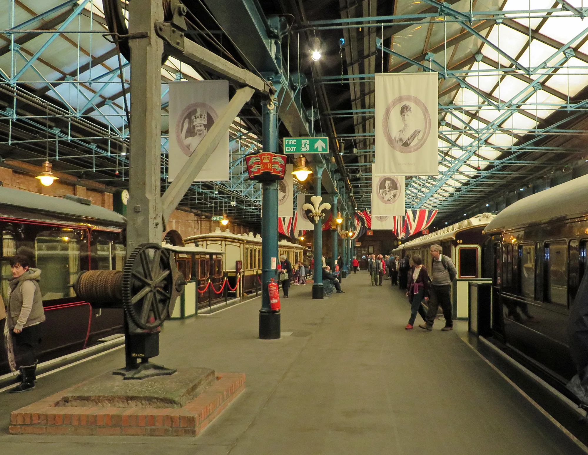Wnętrze hali dworcowej (Station Hall) w National Railway Museum, prezentujące historyczne lokomotywy i wagony po obu stronach szerokiego peronu, z transparentami wiszącymi pod szklanym dachem.