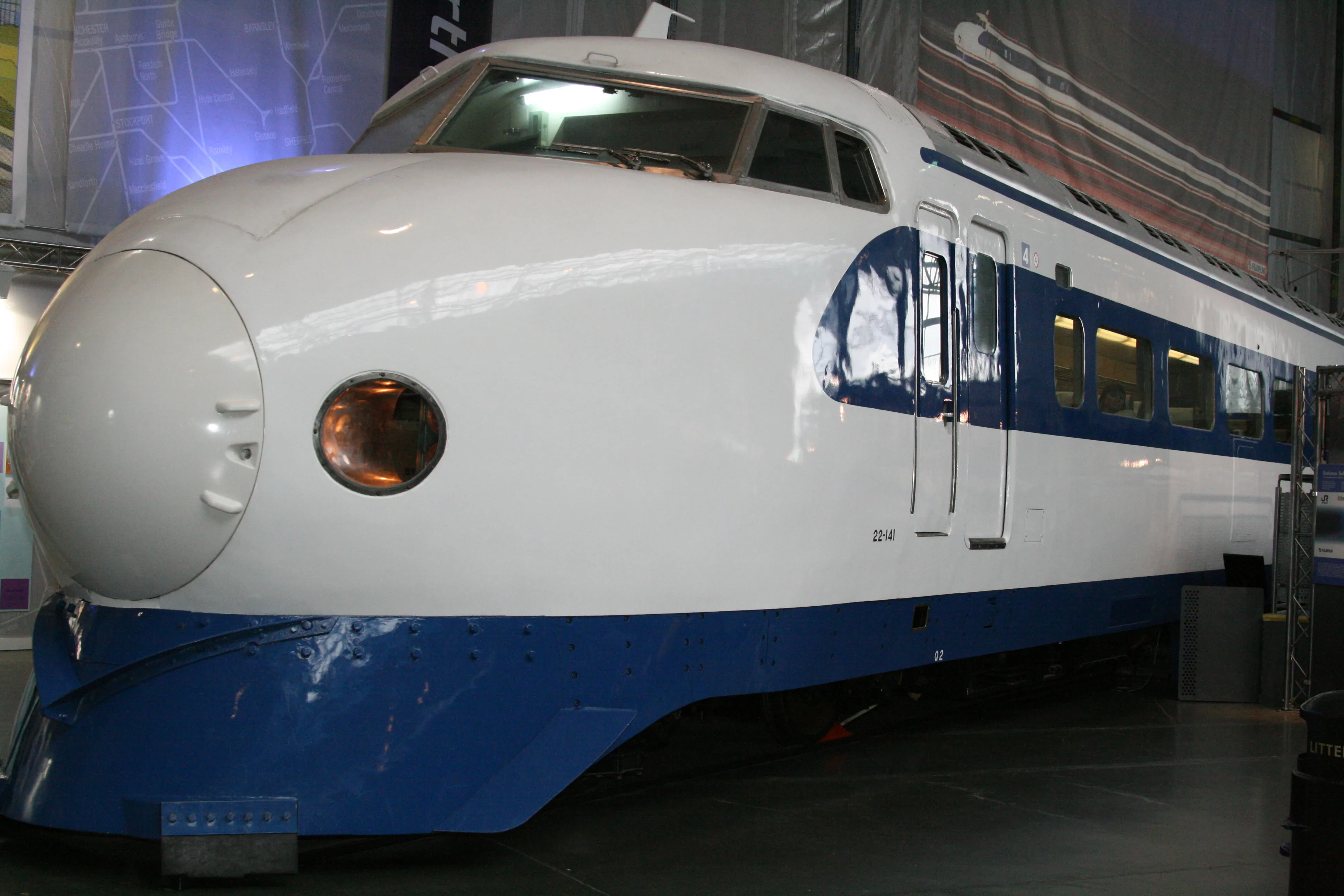 Biało-niebieski pociąg Shinkansen na wystawie wewnątrz budynku, ukazujący zaokrąglony przód, pomarańczowy reflektor i boczne okna pasażerskie