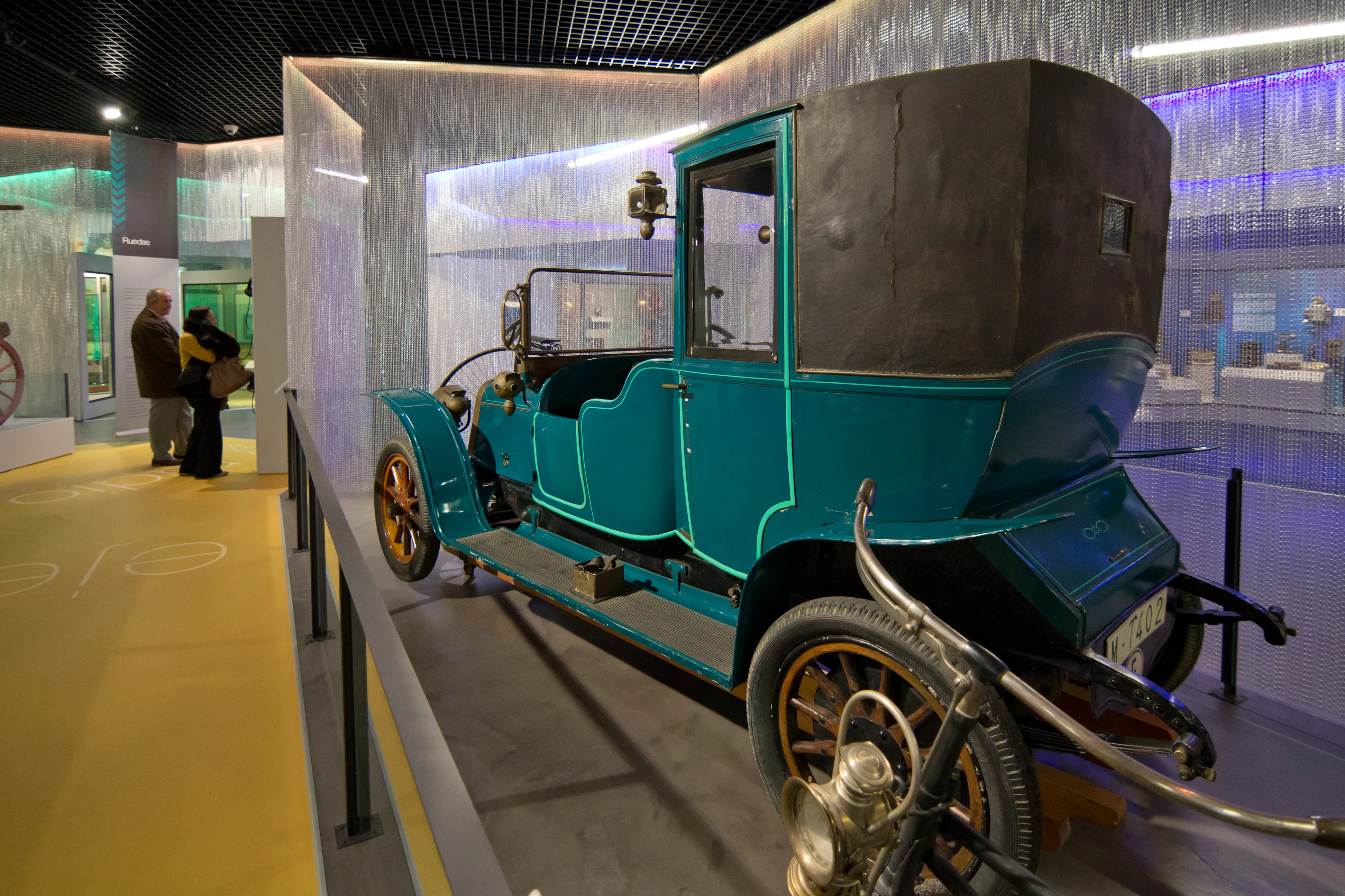 Un automóvil antiguo color turquesa con detalles negros y grandes ruedas de radios, exhibido en una galería de museo con iluminación moderna y barreras de cristal.