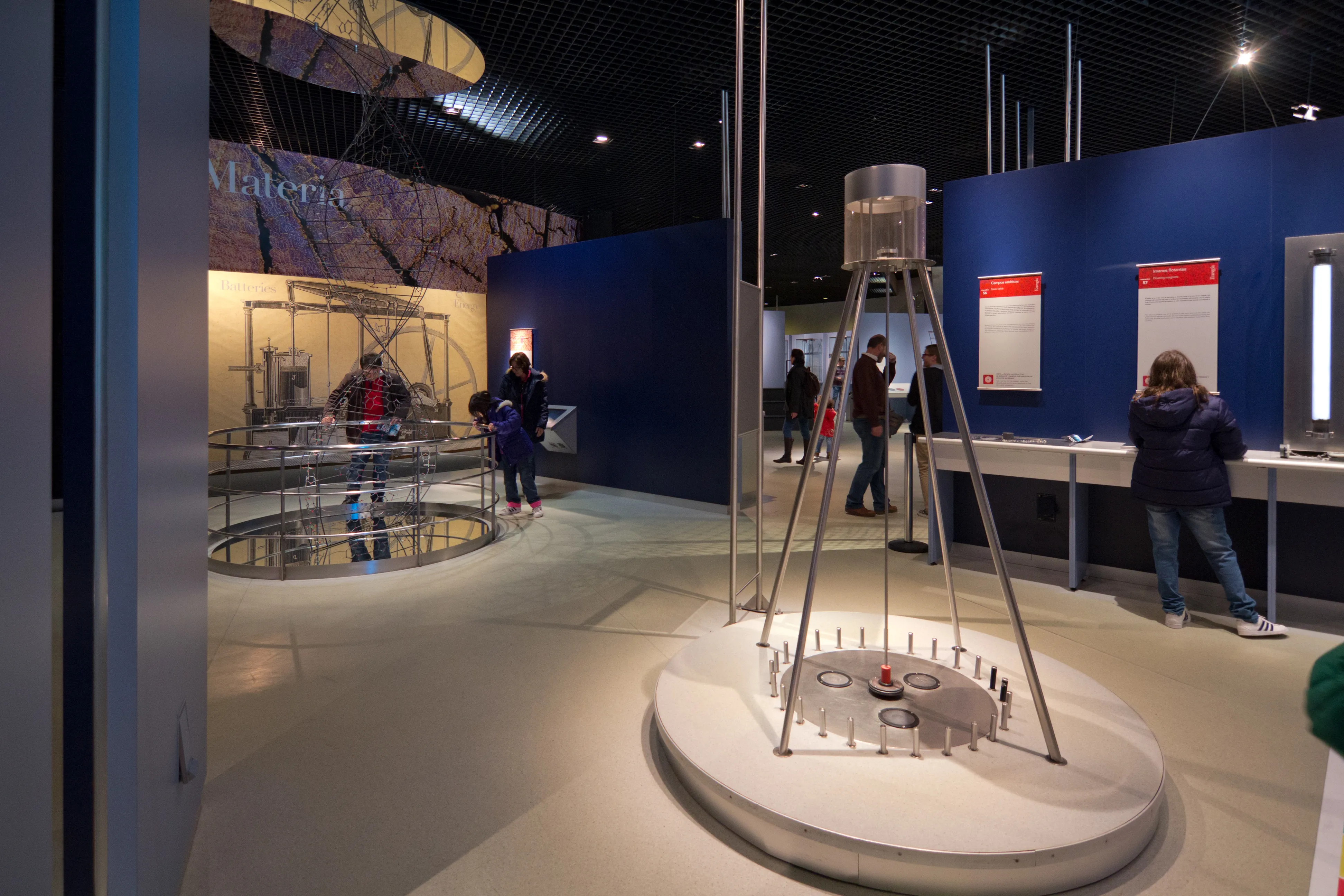 Exhibición interactiva de ciencia con un aparato central sobre una plataforma circular, rodeado de visitantes en una sala de museo con paredes azules y paneles informativos.