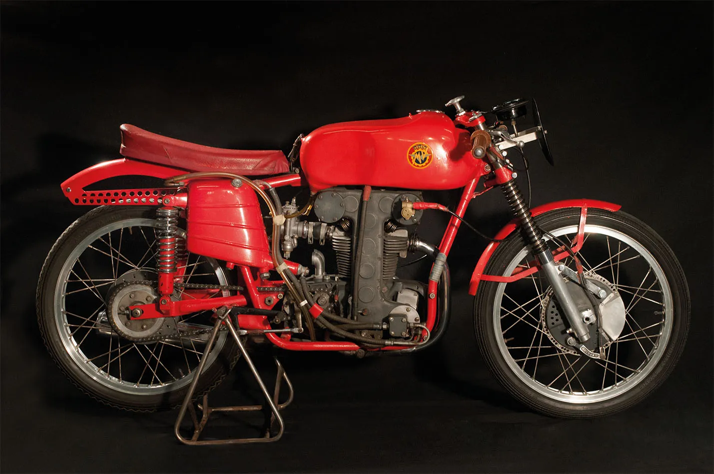 Una motocicleta vintage roja con motor negro y detalles cromados en exhibición sobre un fondo oscuro