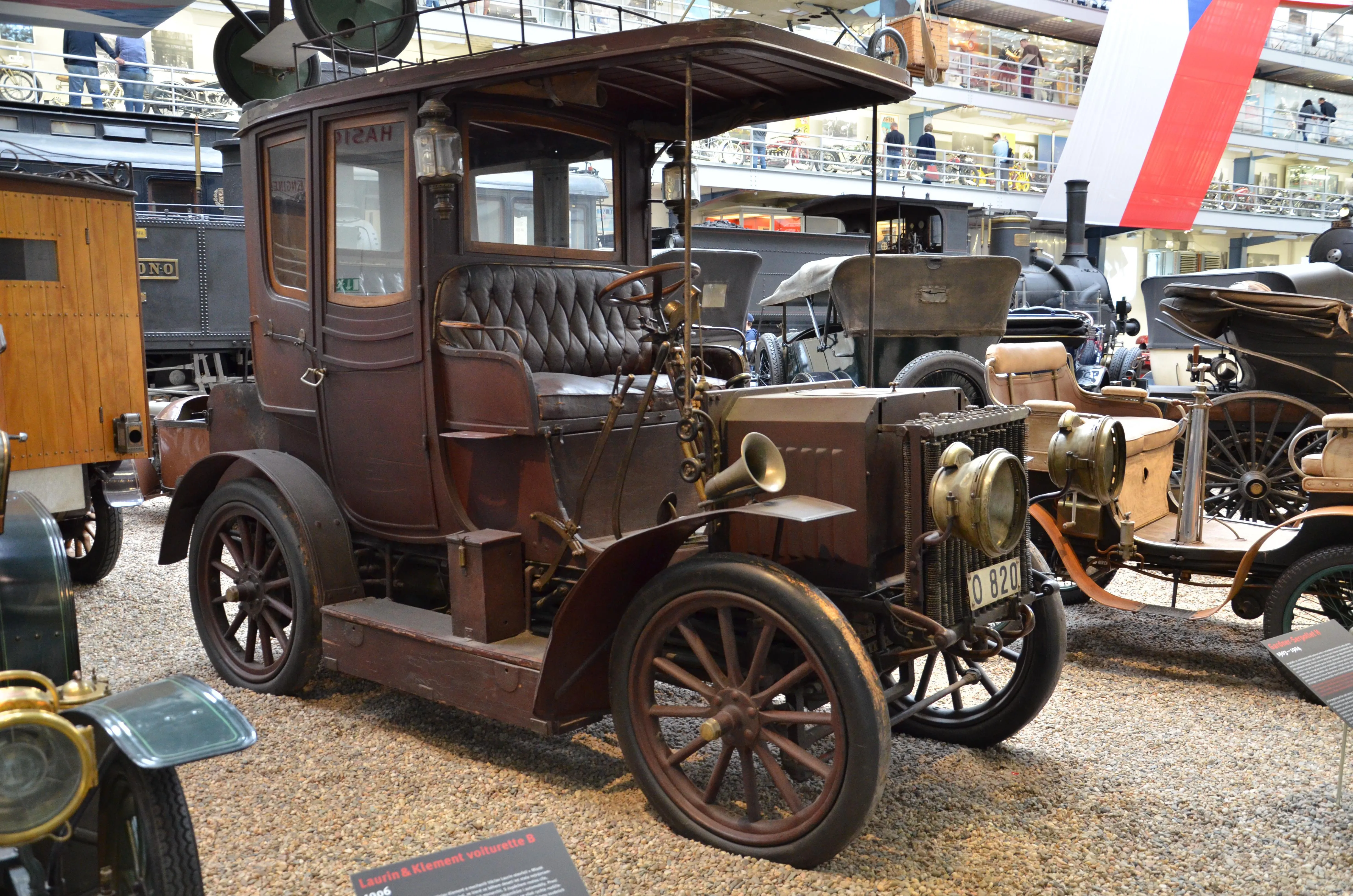A vintage brown automobile with wooden wheels and a leather interior, displayed in a museum hall with other historical vehicles.