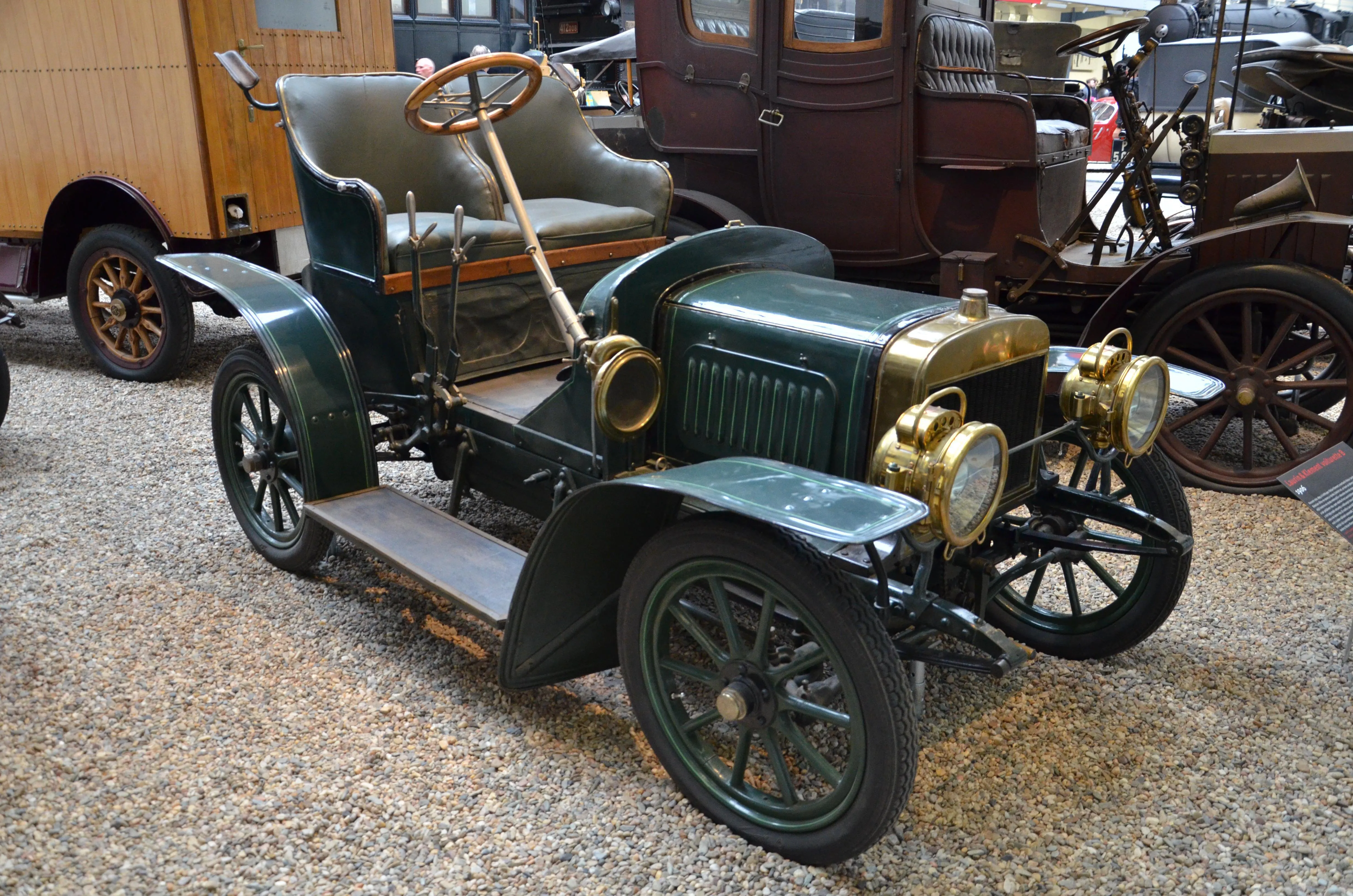 A green vintage automobile with brass headlights and a wooden steering wheel displayed in a museum hall