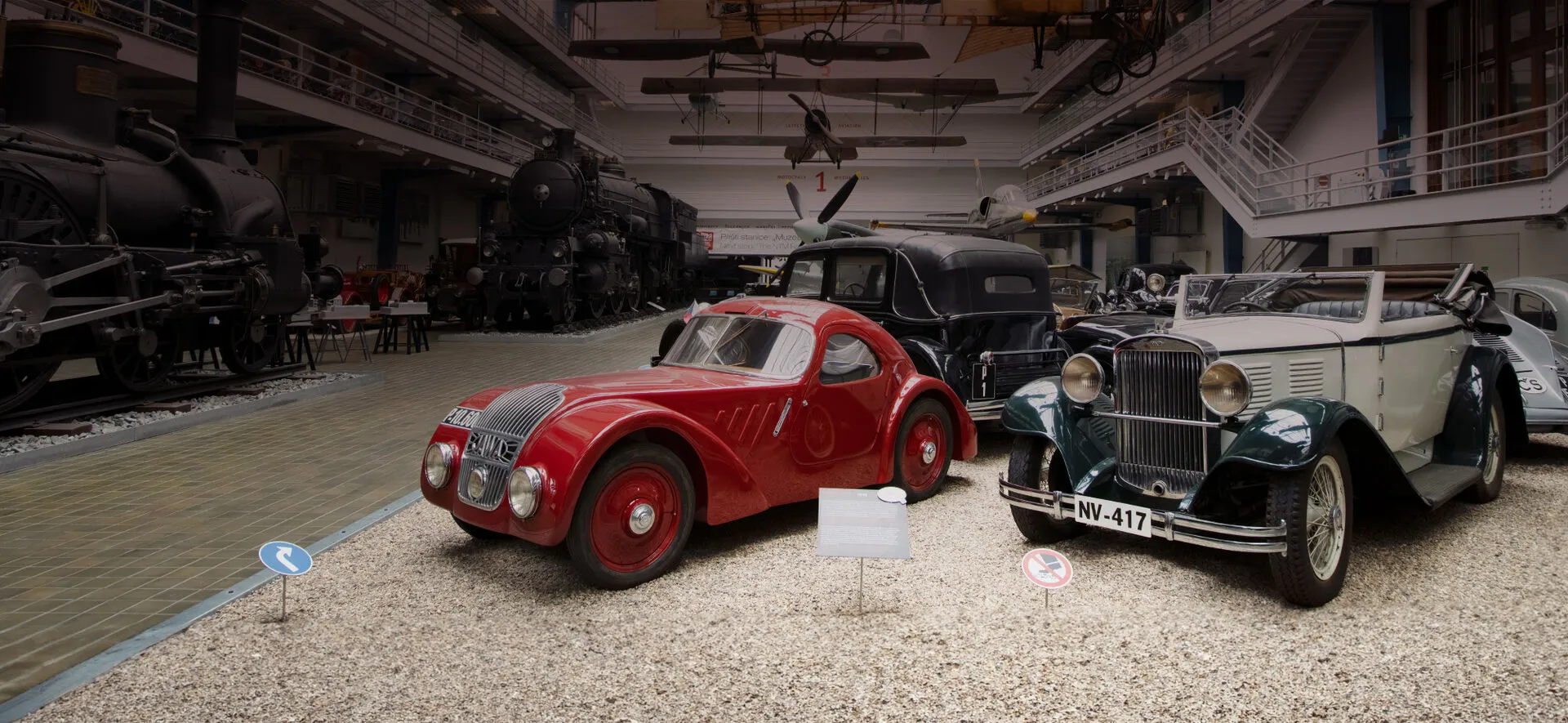 Two vintage cars, one red and one silver, displayed in a large museum hall with a steam locomotive in the background.