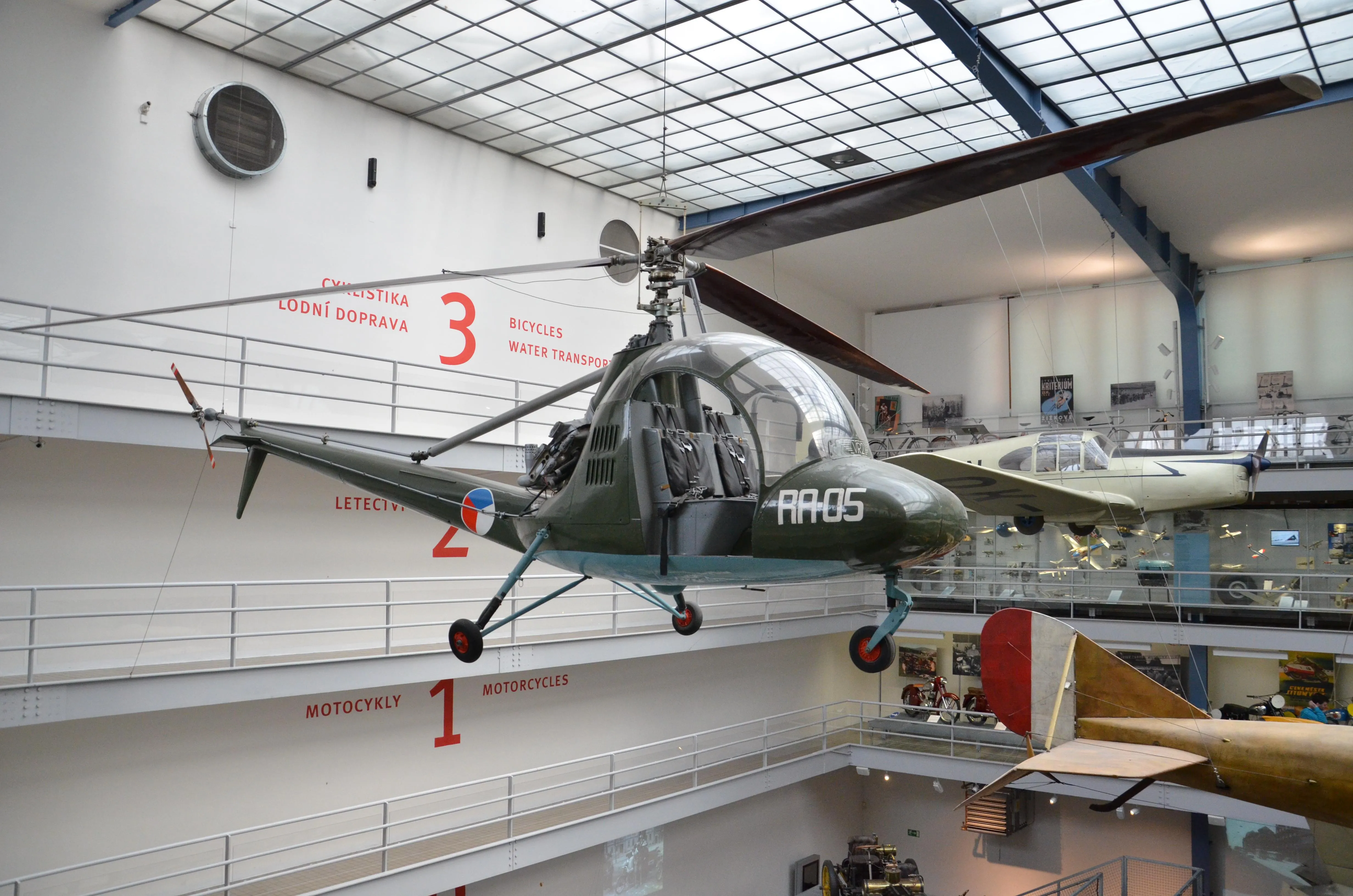 A green helicopter suspended from the ceiling in a museum hall with numbered sections on the wall