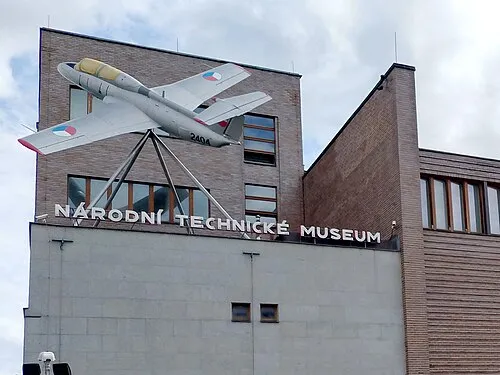 A large silver airplane mounted on a stand in front of a brick building with the sign 'NÁRODNÍ TECHNICKÉ MUSEUM' on the facade.
