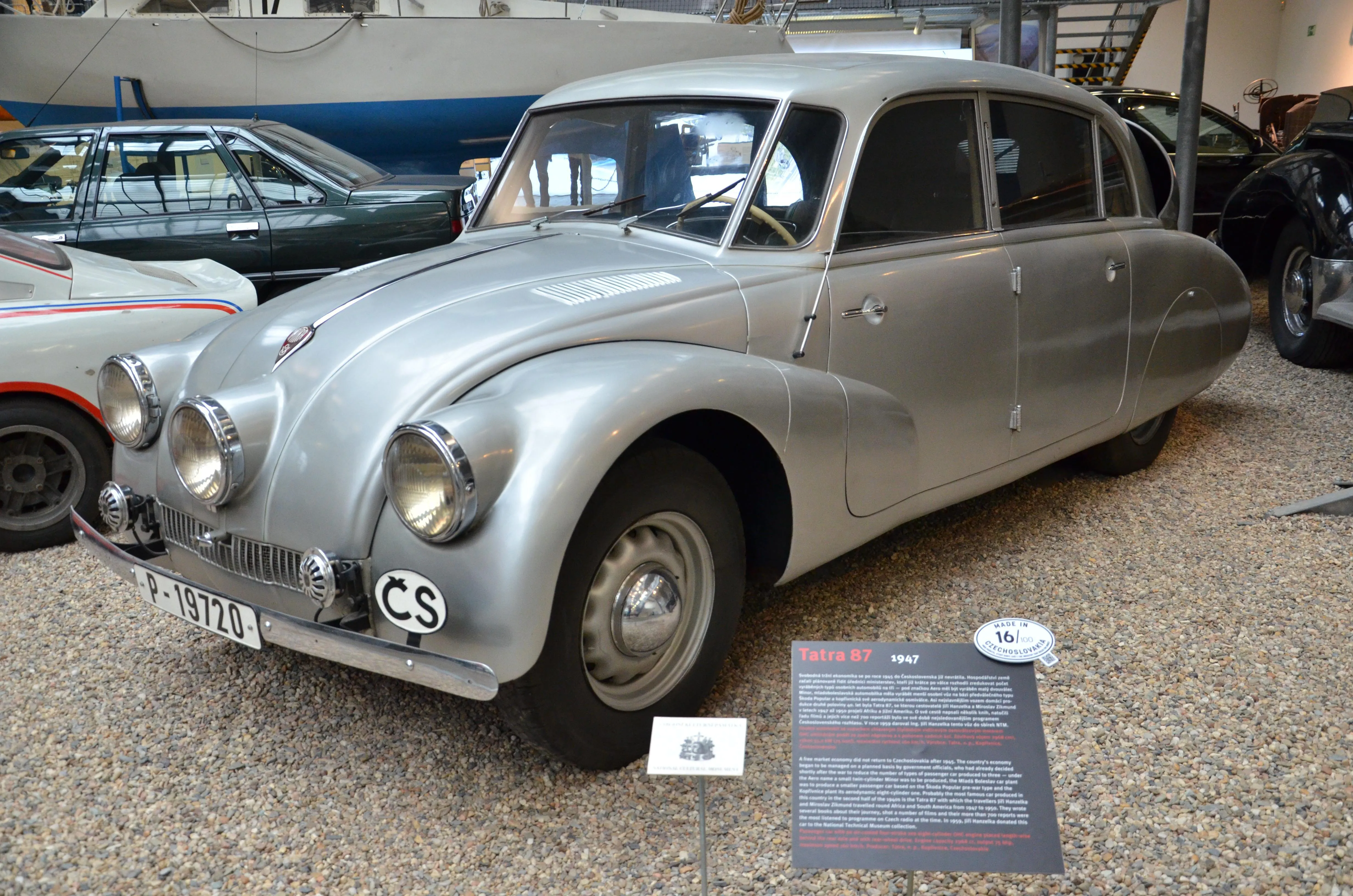 A silver vintage Tatra 87 car with round headlights and a CS license plate, displayed indoors with informational plaques and other cars in the background.